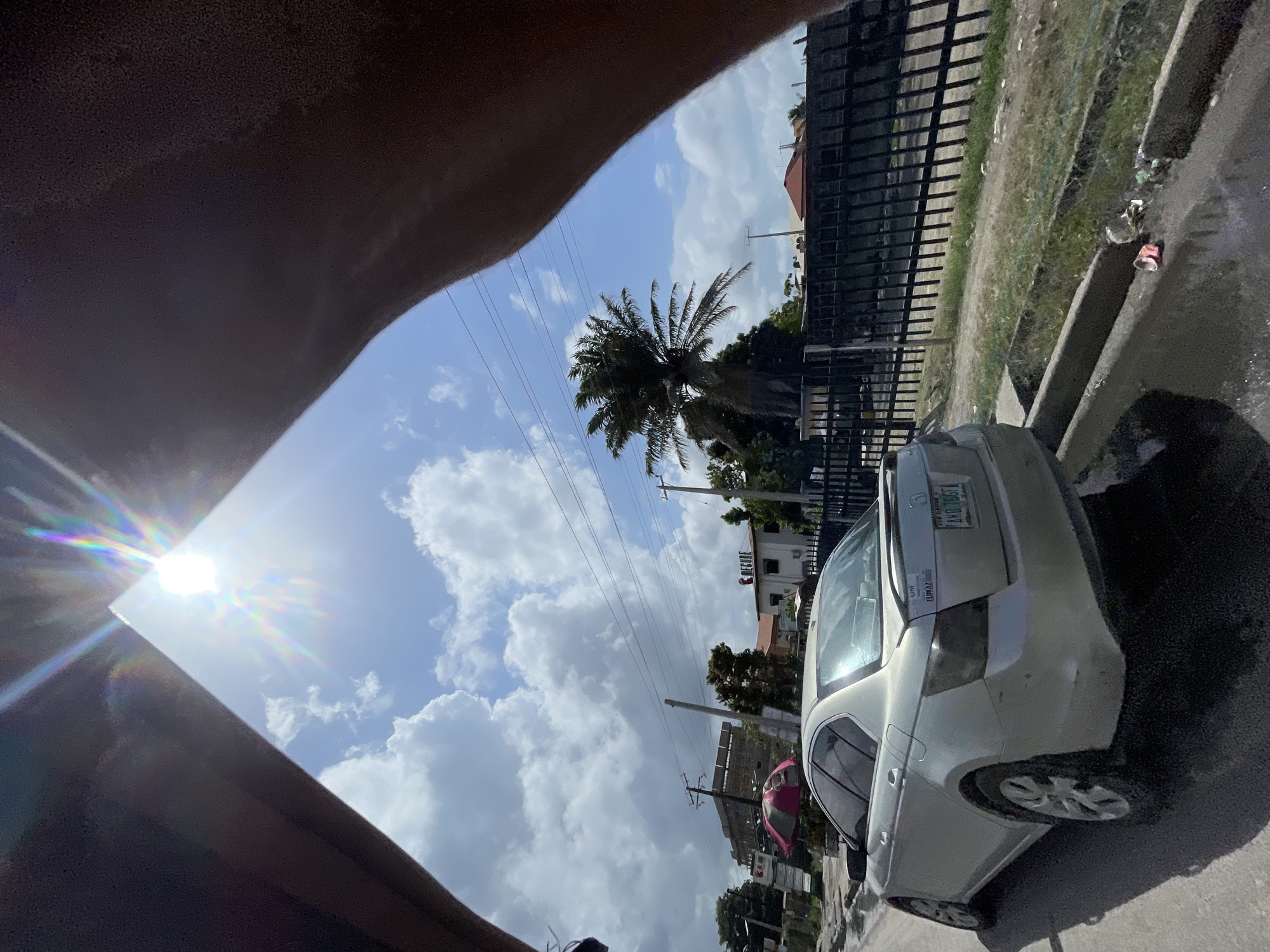 The image shows a view from inside a vehicle, possibly a car. Part of the car's interior is visible in the foreground. The view outside includes a sunny sky with clouds, a palm tree, and utility poles with wires. Adjacent to the car is a fence and some roadside vegetation. Parked next to the road is a silver car. The license plate is partially visible and contains the lettering: "AYD 146GT" and a sticker labeled "UFR". The image was taken during daylight hours in Ado, Nigeria.