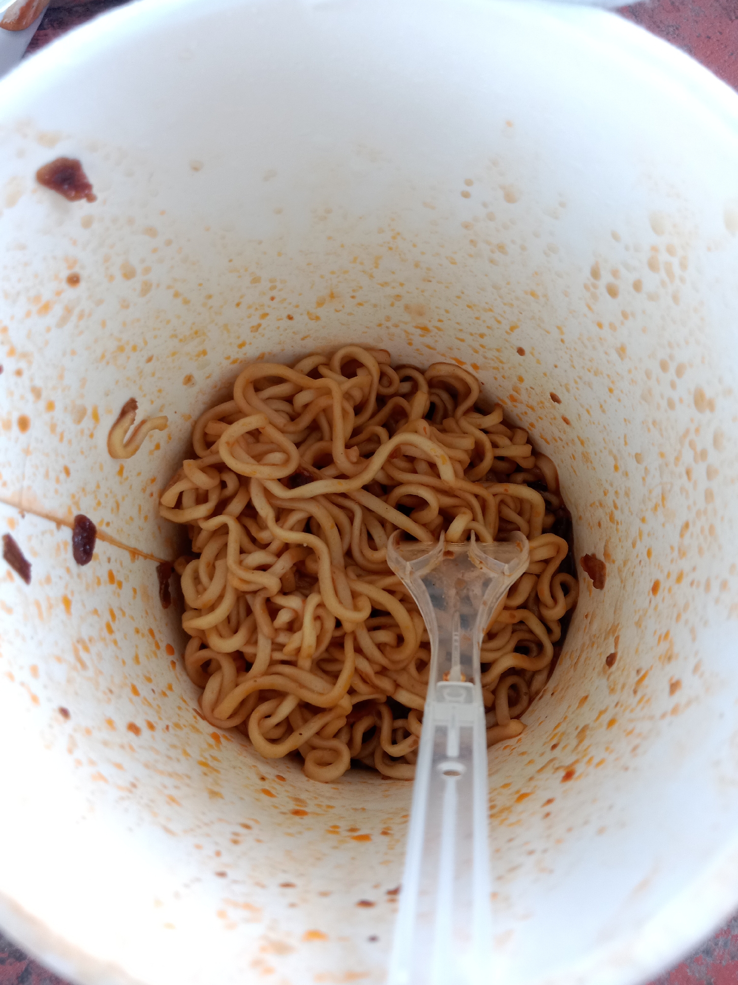 A close-up, top-down shot displays a white cup filled with instant noodles and a clear plastic fork. The inside of the cup is stained with orange and brown splotches from the noodle sauce. The noodles are a light brown color and appear to be coated in a savory sauce. The fork is positioned in the center of the cup, with its prongs submerged in the noodles. The background is blurred, but suggests an outdoor setting. The image is located in Kemamang, Indonesia.