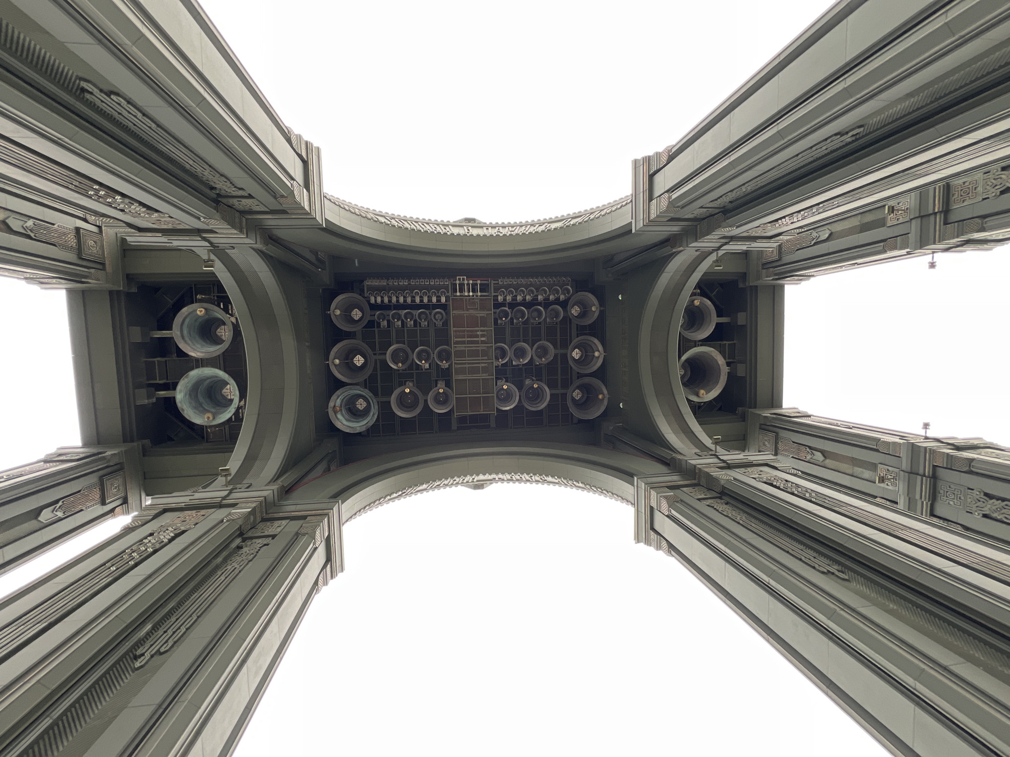 This is a low-angle shot looking up at the bottom of a large structure, likely a bridge or tower, located in Zelenaya Roshcha, Russia. The structure features four massive, parallel, green-colored pillars that seem to support a curved, arched ceiling overhead. On the ceiling, there are many bells arranged in rows, with a ladder visible in the center. The overall architectural style is ornate with decorative elements on the pillars. The background is a uniformly bright white, suggesting a cloudy sky or intentionally overexposed photography.