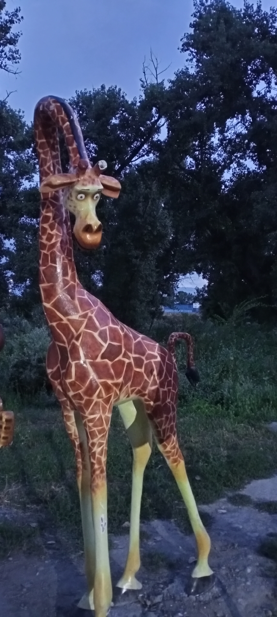 The image captures a statue of a giraffe with a cartoonish appearance, featuring wide eyes and a distinctive facial expression. The statue is positioned outdoors, with a backdrop of lush green foliage and dark trees against a twilight sky. The giraffe is adorned with its characteristic brown and beige patterned coat, and its legs transition to a pale yellow and green hue near the hooves. The statue is standing on what appears to be a path or ground composed of gravel and darker material. In the background, beyond the trees, there are faint hints of distant lights and structures, possibly indicating a town or settlement. The overall lighting suggests it is dusk or evening. The location is identified as Sokuluk, Kyrgyzstan.