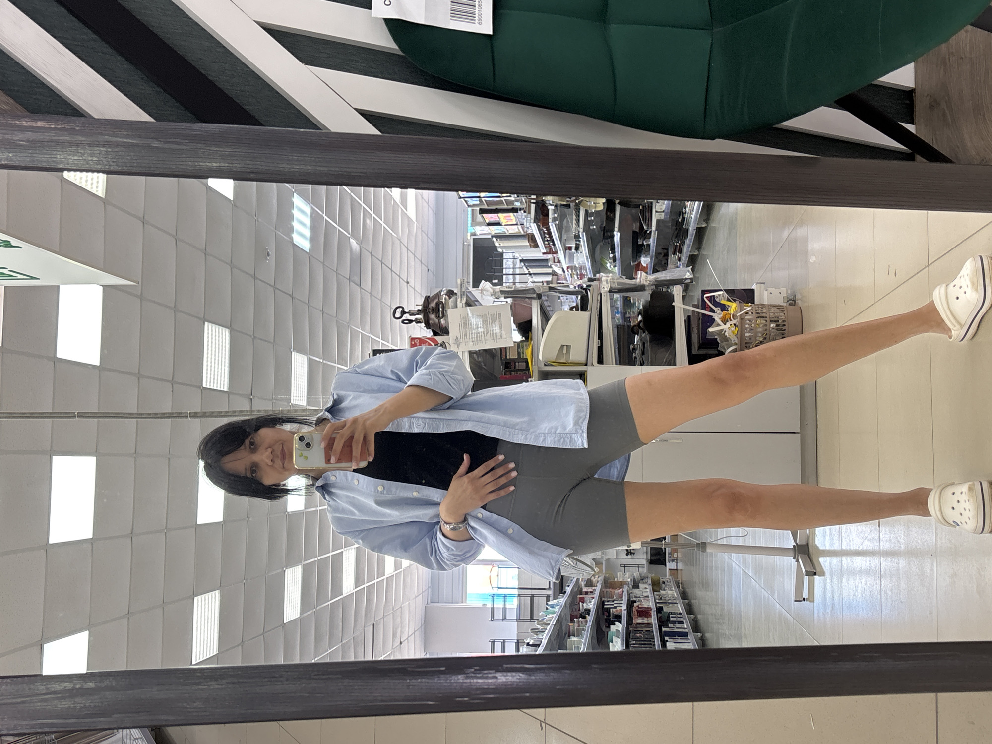 A woman is standing in front of a mirror, taking a selfie in a retail store. She is wearing a light blue button-down shirt, a black tank top, grey biker shorts, and white Crocs. Her left hand is resting on her stomach, and she is holding her phone in her right hand. The background of the image, as seen in the mirror's reflection, shows aisles of merchandise, fluorescent lighting on the ceiling, and a patterned decorative element on the wall. The location is identified as a retail store in Chaikovsky, Russia.
