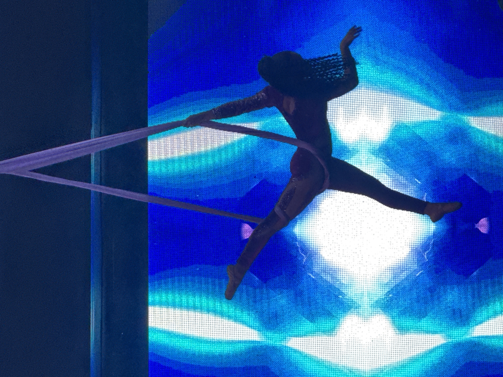 A lone aerialist performs a routine on a suspended fabric against a vibrant blue and white LED screen backdrop. The aerialist, clad in a sparkling, form-fitting outfit, is positioned in mid-air, suspended by two pale pink fabric silks. Their body is arched, with one leg extended straight upwards and the other bent at the knee. The aerialist's arms are in dynamic poses, one reaching upwards along the silk and the other extended sideways. The LED screen displays an abstract, luminous pattern of blues and whites, creating a futuristic and theatrical atmosphere. The scene appears to be indoors, possibly in a performance venue or theater. The image captures a moment of intense physicality and artistic expression. The location in Manavgat, Turkey is not discernible from the image itself.
