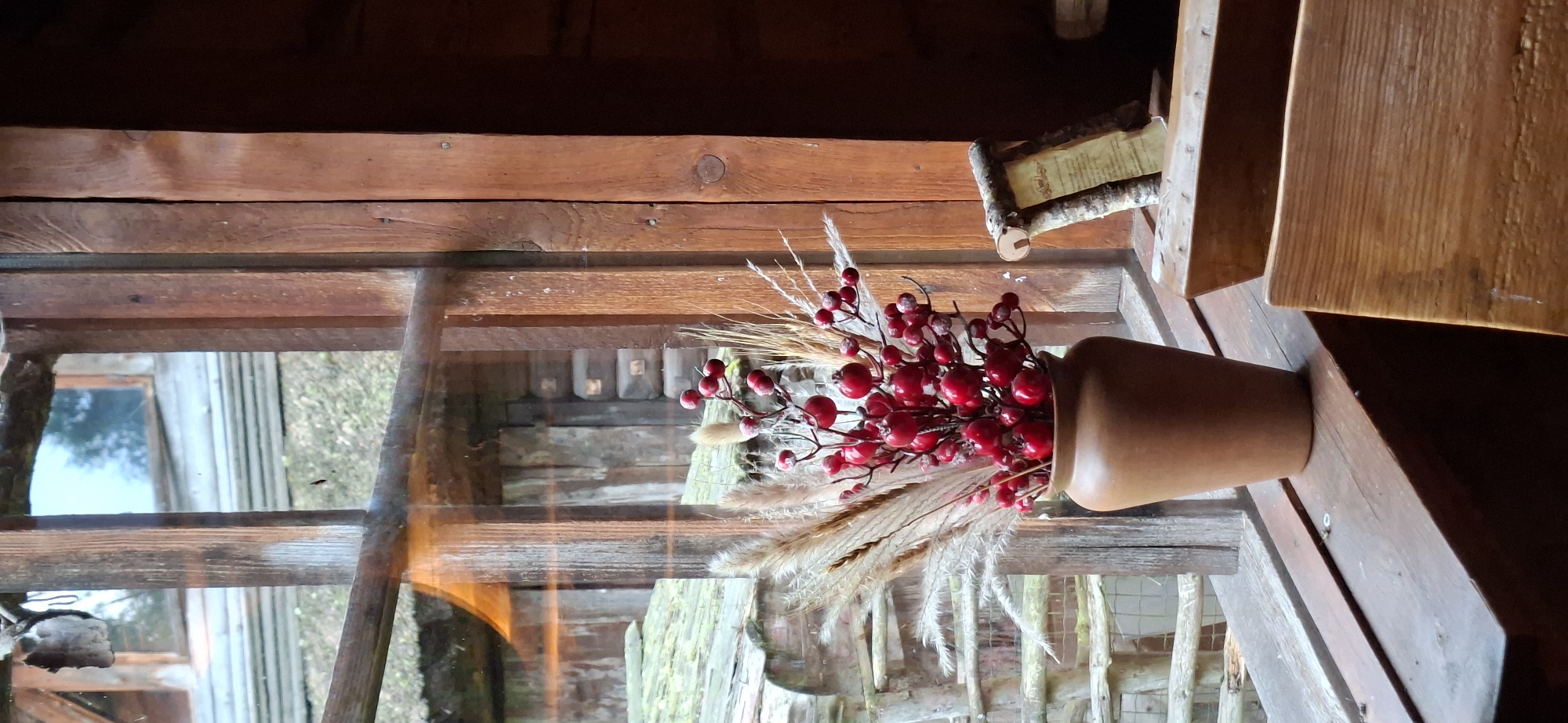 The image depicts a rustic interior scene, likely within a wooden structure, with a window as the central focus. A vase filled with red berries and dried grasses sits on the wooden windowsill. Through the window, a glimpse of an outdoor environment is visible, featuring wooden buildings, a thatched roof, and a wooden staircase. The overall aesthetic suggests a rural or historical setting. The scene is devoid of any people or discernible actions. The lighting appears to be natural, filtering in from the window, creating a slightly diffused and warm atmosphere. The image was taken in Saula, Estonia, within a locality characterized by its traditional architecture and natural surroundings.
