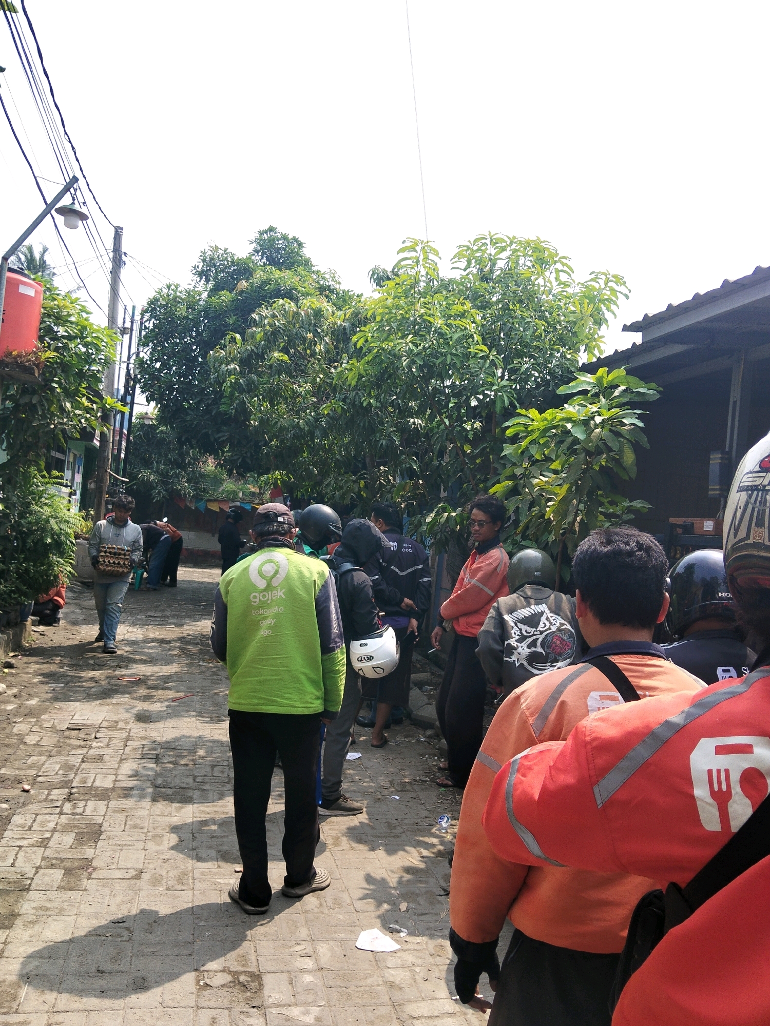 In the city of Rajegmulya, Indonesia, a group of people, primarily men, are gathered on a brick-paved alleyway lined with trees and buildings. A significant portion of the individuals appear to be delivery drivers, indicated by their uniforms and equipment. One man in the foreground wears a bright green jacket with the "Gojek" logo, a prominent Indonesian ride-hailing and delivery service. To his left, another man is seen carrying a crate of what appears to be eggs. Several individuals are wearing helmets, suggesting they are on motorcycles, some of which are partially visible. The atmosphere suggests a waiting or gathering point, possibly for assignments or a break. The environment includes overhead power lines and the natural elements of trees, casting shadows on the ground.