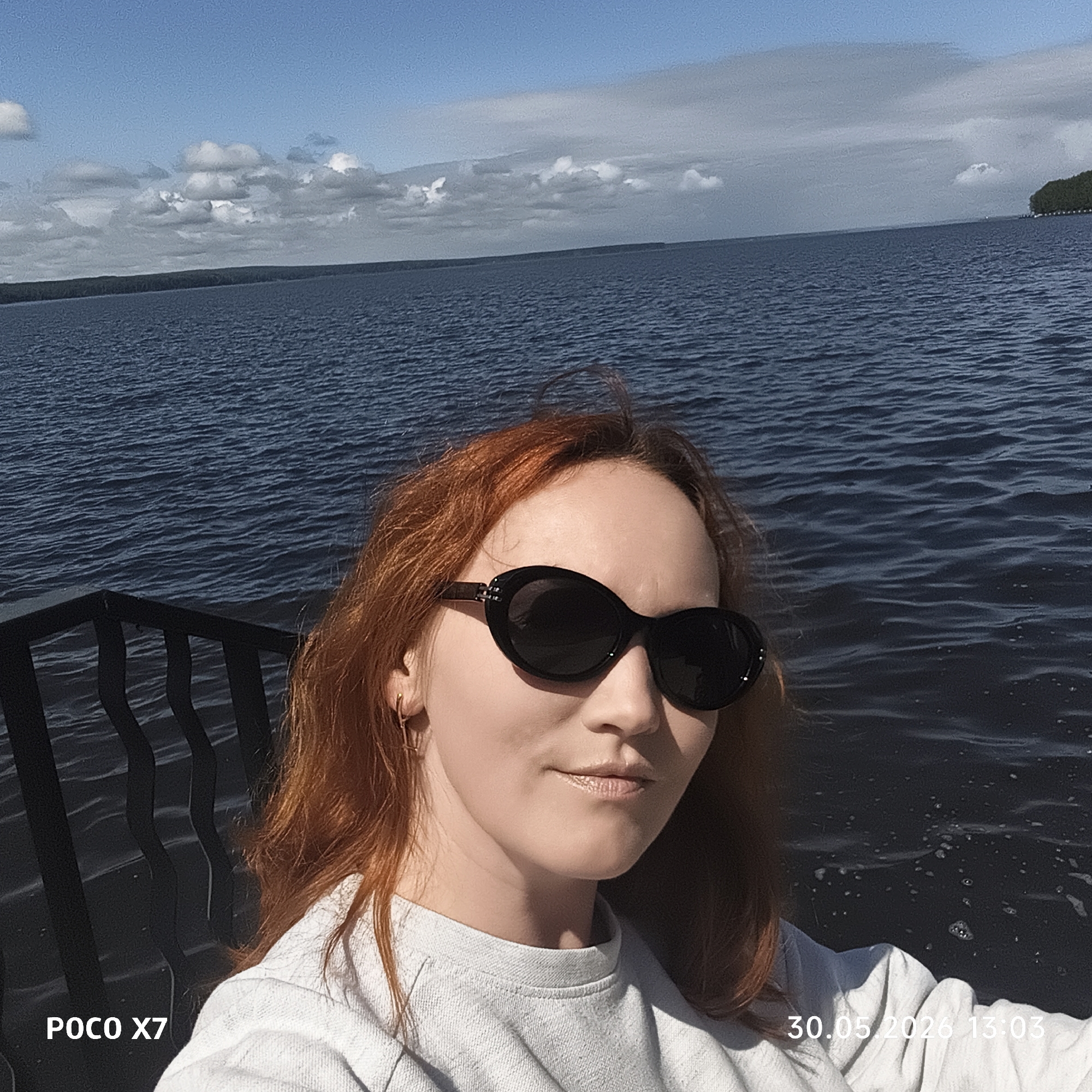 A selfie-style image features a fair-skinned woman with reddish-orange hair wearing large, black, oval sunglasses and a light grey sweatshirt. She is positioned against a background of a large body of water, possibly a lake or reservoir, with visible waves and a distant shoreline covered in trees. The sky is partly cloudy. The left side of the frame includes a section of black wrought-iron railing. Text at the bottom left corner reads "POCO X7," indicating the device used to take the photo, and at the bottom right corner, "30.05.2026 13:03" provides a timestamp. Given the city of Chaikovsky, Russia, is specified, the water body is likely the Votkinsk Reservoir. The woman is likely posing for the photo, and the environment suggests an outdoor location near the water's edge, potentially a pier or viewing platform.