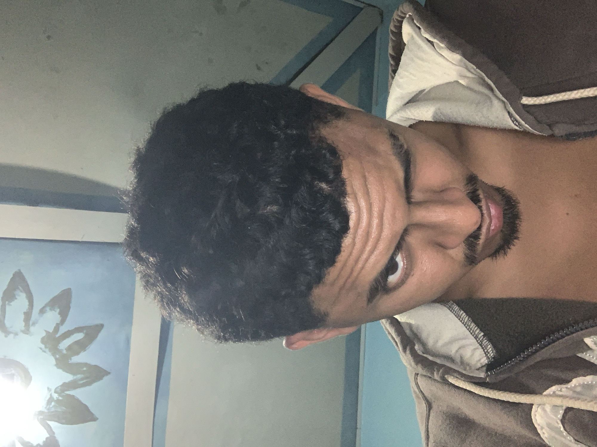 The image shows a close-up, overhead shot of a young man with dark, curly hair and a goatee. He is wearing a brown hooded sweatshirt with a cream-colored lining and a zipper. His left eye is wide open and looking directly at the camera, while his right eye is squinted as if winking. His forehead is furrowed, and his mouth is slightly open. In the background, there appears to be a light blue ceiling with a painted white sun or flower design and some blue striped accents. The lighting suggests an indoor environment. The image does not provide any visual cues or context that would allow for the determination of its location within Santa Ana, Venezuela.