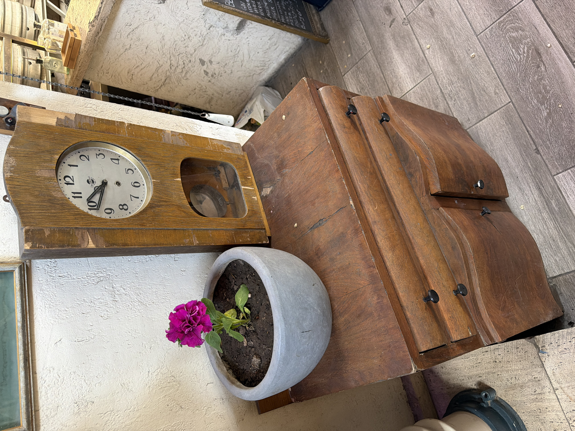 Here is a description of the image.
The image presents a cluttered indoor scene, possibly within a café or shop in Yerevan, Armenia, judging from the ambiance and decor.
A wooden clock with a circular face showing the time to be approximately 10:35 hangs slightly off-center. Beside it is a rounded grey flower pot, containing soil and a vibrant purple flower with green leaves. The flower pot sits on a brown wooden cabinet with arched drawers. In the background, partially visible is a chalkboard menu. The flooring appears to be made of grey wooden planks.
The overall atmosphere is one of organized clutter with a somewhat rustic and lived-in aesthetic.