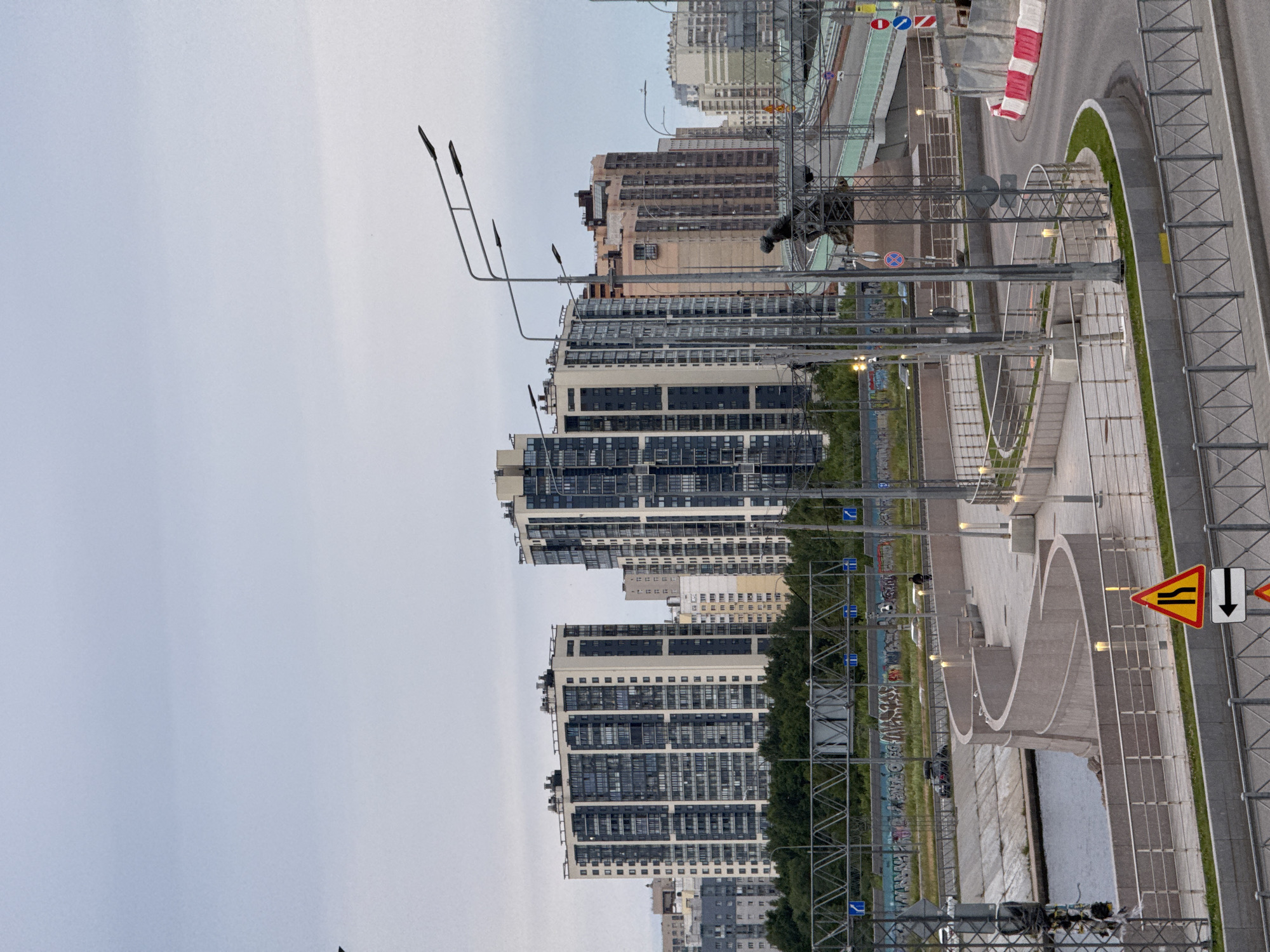 This is an eye-level, wide shot of a modern urban landscape in Saint Petersburg, Russia. The image is dominated by several tall, contemporary apartment buildings with distinct architectural features, including balconies and large windows. In the foreground, a metal fence runs along a raised walkway or promenade, which curves gently. This walkway is bordered by a grassy area and a body of water on one side, and a road on the other. The road itself features traffic signs, including a warning sign indicating a narrowing of the road ahead with a downward arrow, and a circular sign with a red border and a diagonal red bar. A red and white striped barrier is also visible on the side of the road.
Several streetlights are present, suggesting it is either dusk or an overcast day. The sky is a pale, uniform grey, indicating cloudy or hazy weather. The overall mood is one of quiet urban development. There are no people clearly visible in the image, but there are some parked cars. The architectural style of the buildings and the presence of specific traffic signs hint at a modern, planned urban area within the city. The image is oriented vertically. No discernible text is visible on the buildings or other prominent structures.