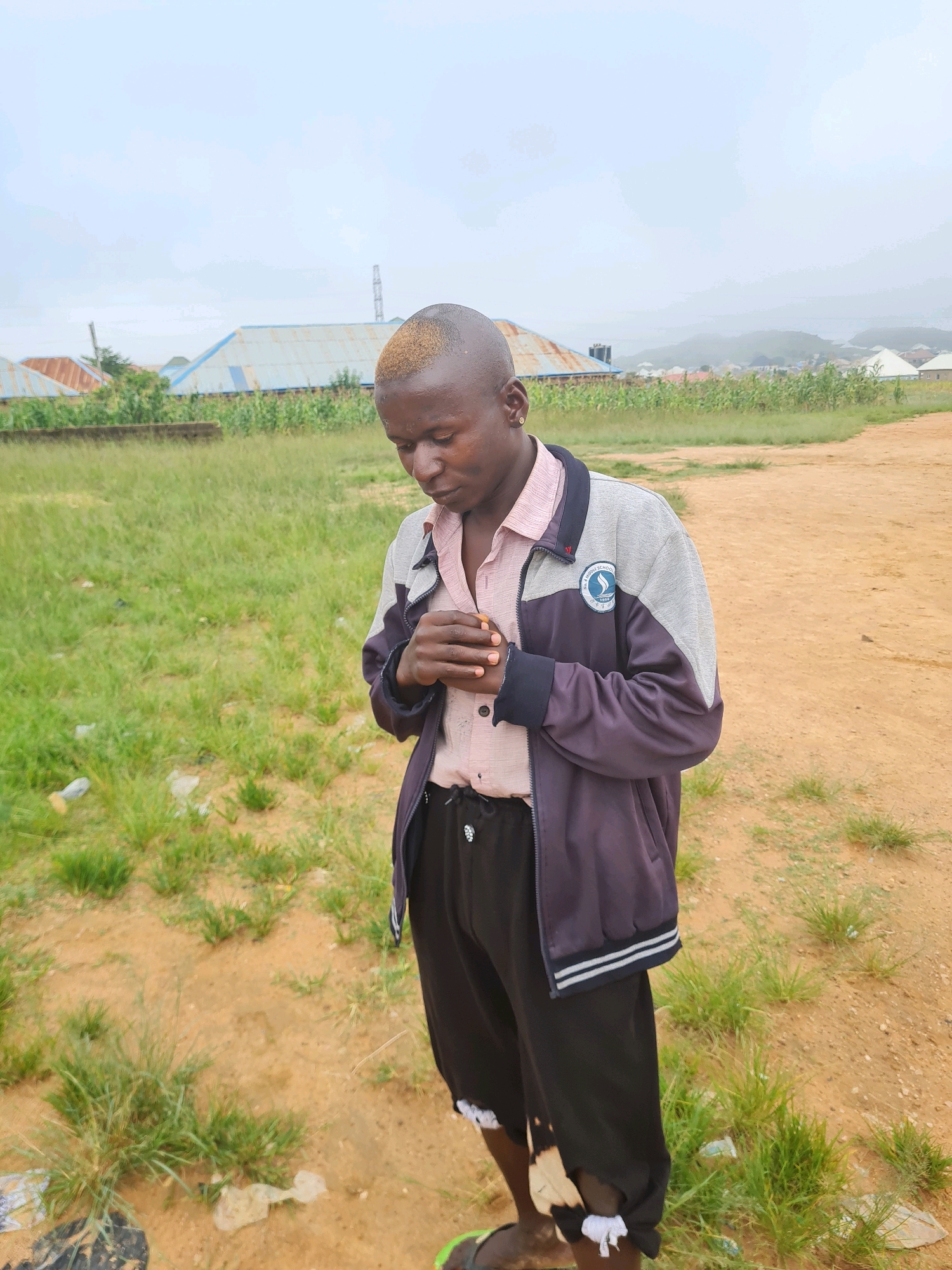 A young person with a shaved head dyed blond stands outdoors in a dusty, unpaved area. They are wearing a light pink collared shirt and a dark blue and gray jacket with a school emblem. Black pants with frayed edges and white ties are also visible. The background shows a grassy field, some buildings with corrugated metal roofs, and a distant hazy cityscape with hills, suggesting an urban or semi-urban environment in Jos, Nigeria. The sky is overcast, and there is some litter visible in the foreground. The person is looking down, with their hands clasped in front of their chest.