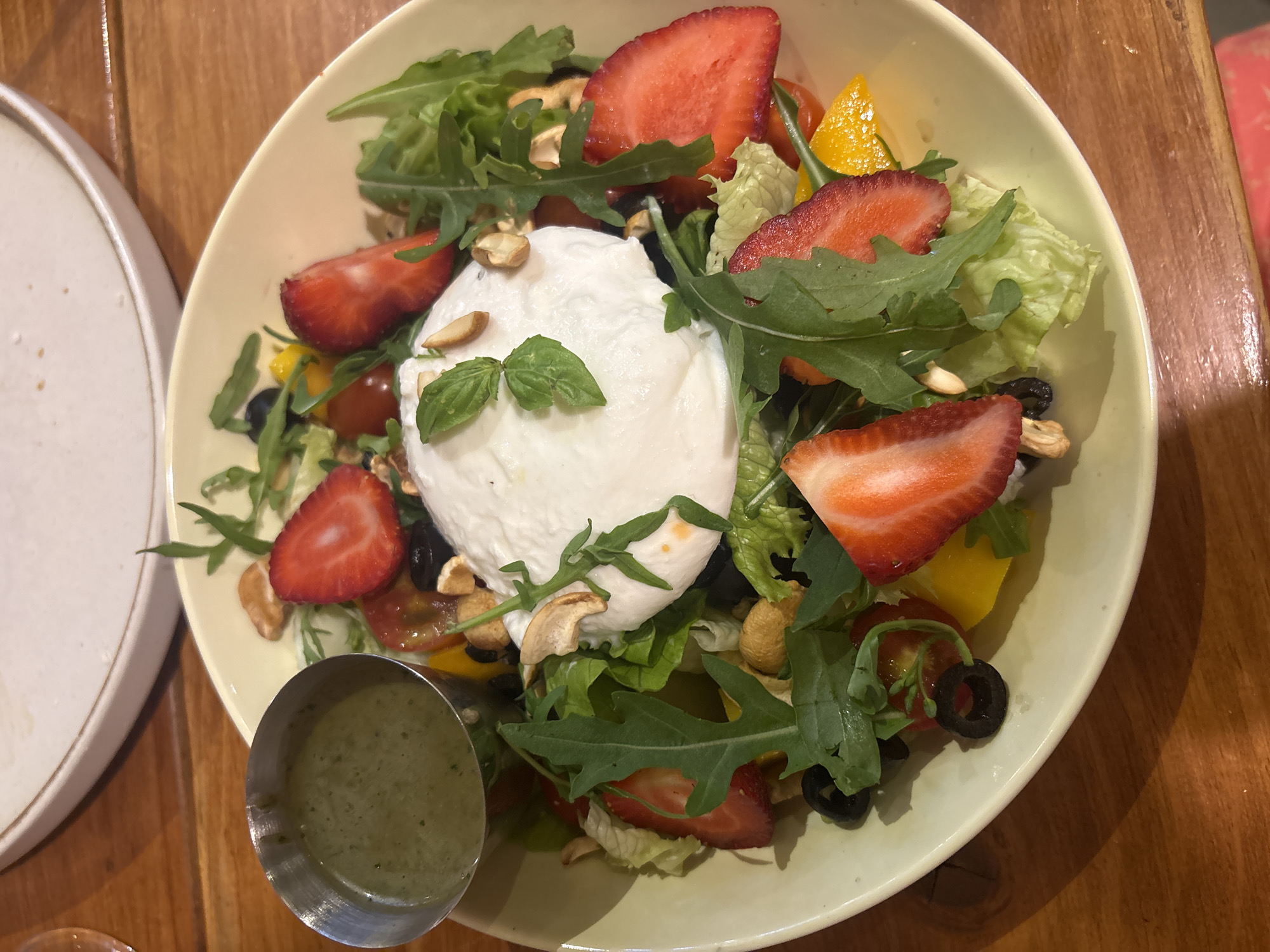 A top-down, medium shot captures a vibrant salad served in a pale yellow bowl, positioned on a wooden table. The salad is a colorful medley of ingredients, including fresh green lettuce, sliced strawberries, cherry tomatoes, black olives, yellow bell pepper chunks, cashews, and a generous dollop of creamy white burrata cheese at its center, garnished with basil leaves. A small metal cup filled with green dressing sits to the left of the bowl. The background is slightly blurred, showing a white plate and a portion of the wooden table surface. The overall scene suggests a meal at a restaurant in Da Nang, Vietnam, given the context provided.