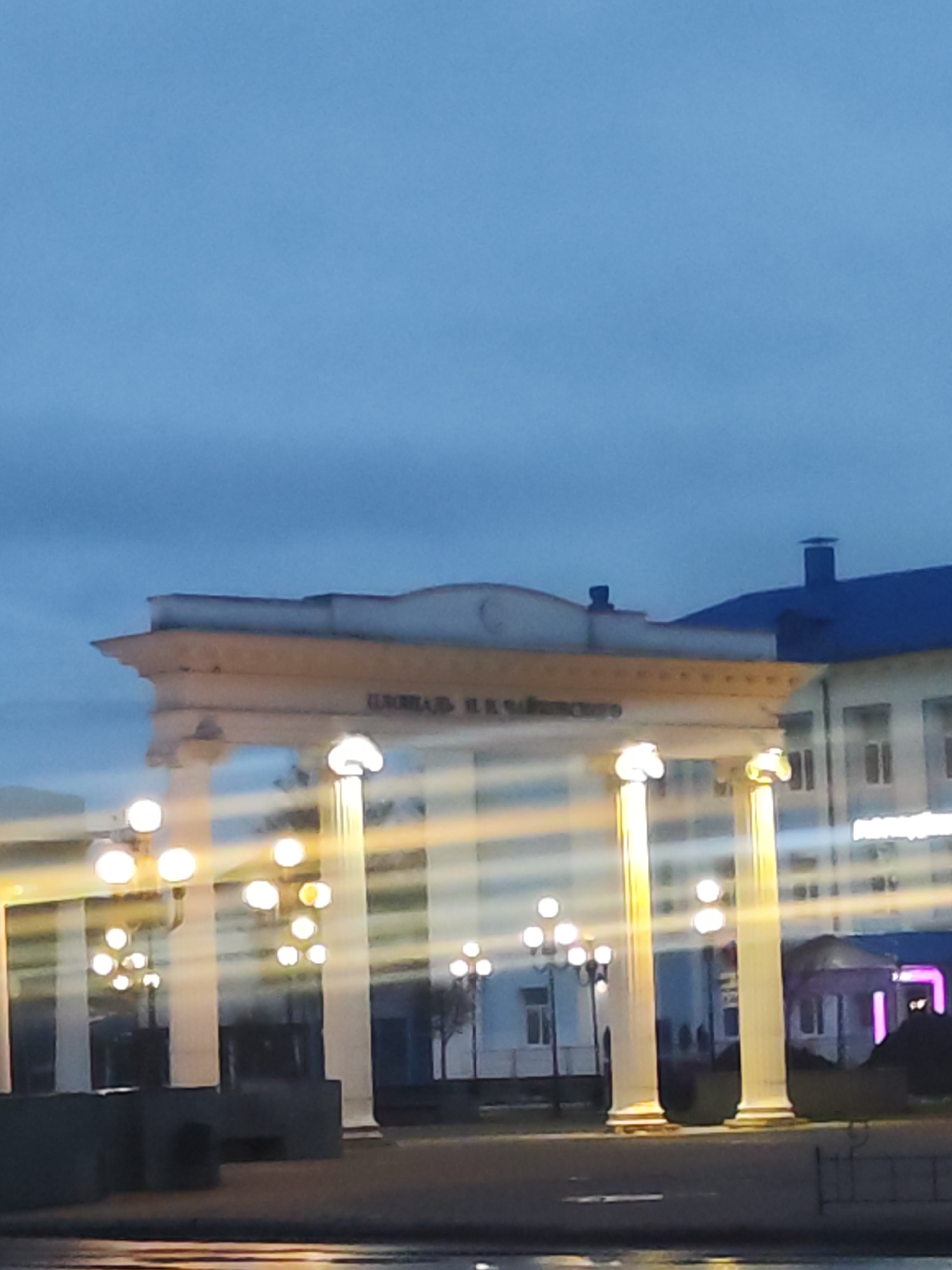 This image shows an architectural structure in Chaikovsky, Russia, at twilight. The structure consists of a horizontal archway supported by several white columns, with glowing spherical lamps atop each column. Inscribed on the archway are Cyrillic characters; the text reads "ШКОЛА №. Я.Н.ЗАЙЦЕВО". Behind the archway, a light-colored building with a dark blue roof can be seen. Additional lampposts are visible in the background, lining what appears to be a street or public space. The foreground has a blurry, undefined texture, potentially water or a reflective surface. No people are clearly visible.