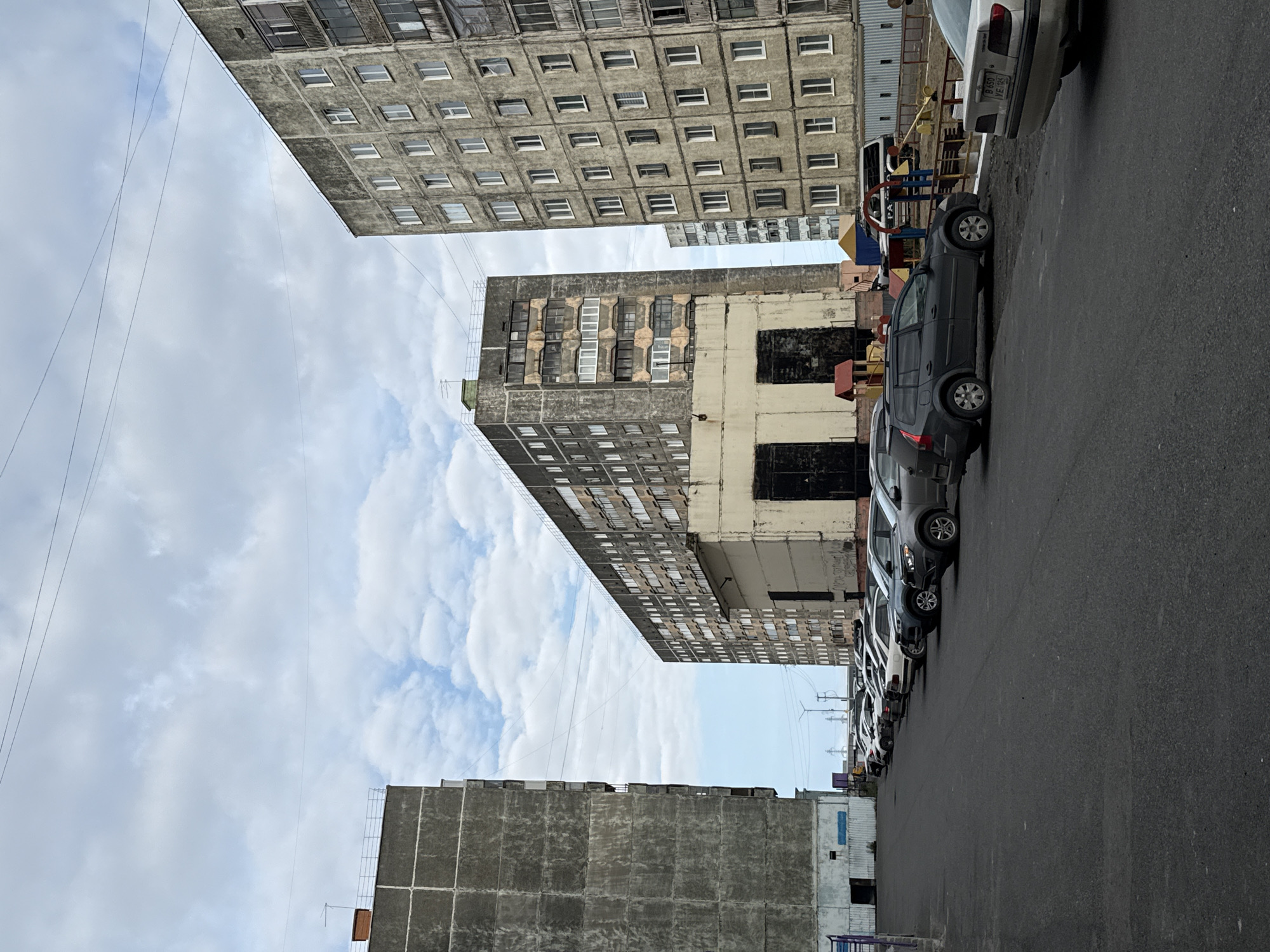 The image depicts a parking lot in Norilsk, Russia, with several apartment buildings in the background. The apartment buildings are tall, concrete structures typical of Soviet-era architecture, with many windows and balconies. The parking lot is paved with asphalt and filled with a row of parked cars, mostly sedans and SUVs. In the middle ground, there is a playground with a slide and swings. The sky is overcast with scattered clouds. The overall scene suggests a typical residential area in a northern city.