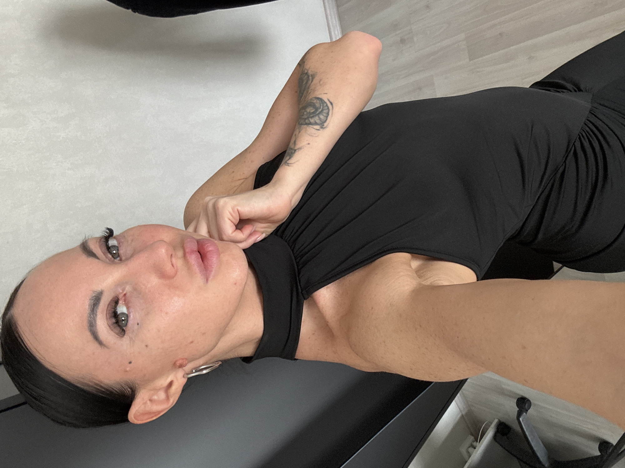 A woman is facing the camera and is angled slightly towards the left. The woman is fair-skinned with dark hair and has dark eyeliner. She is wearing a black top that has a high neckline. There is a tattoo on her left forearm. She is wearing a hoop earring. A black surface is under her left cheek. A wheel from an office chair is in the bottom right corner. The wall behind her is white. The location of Kazan, Russia is not discernible from the image.