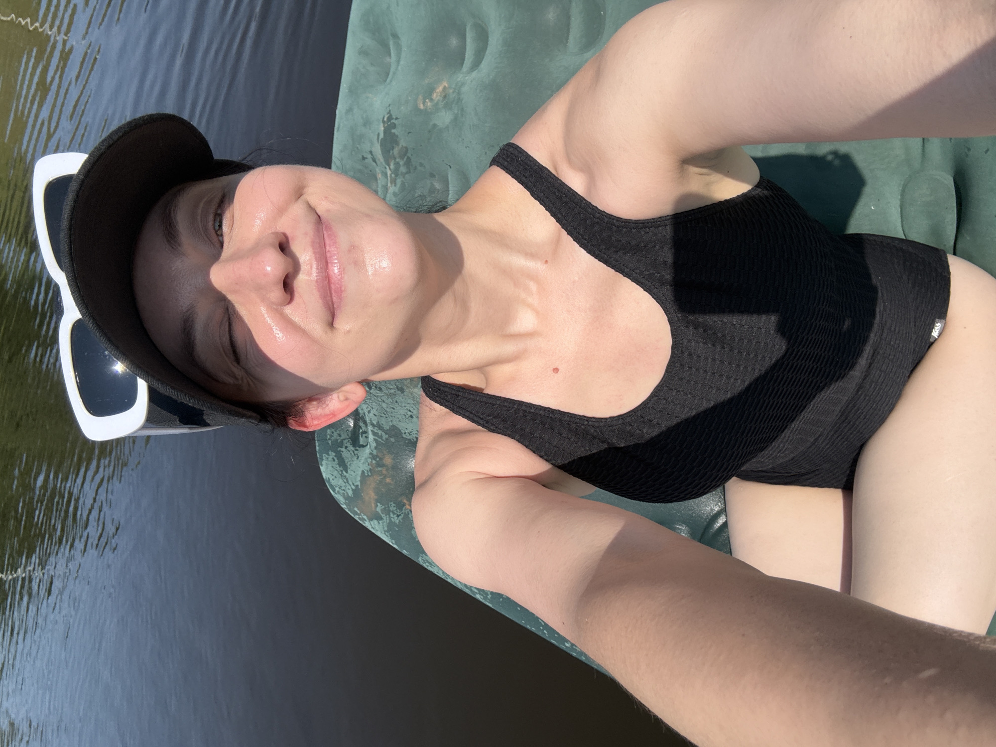 A woman in a black swimsuit and a black baseball cap with white sunglasses on top is floating on an inflatable raft in dark water. She is taking a selfie with a slight smile and one eye closed. The water appears to be a lake or river, with sunlight reflecting off the surface. The raft is green and appears to be partially submerged. The background shows the darker water and a hint of a shoreline or trees on the far side of the water, suggesting an outdoor, possibly rural, setting. The location is not identifiable as Zasechnyi, Russia, as there are no specific landmarks or contextual clues to indicate this.