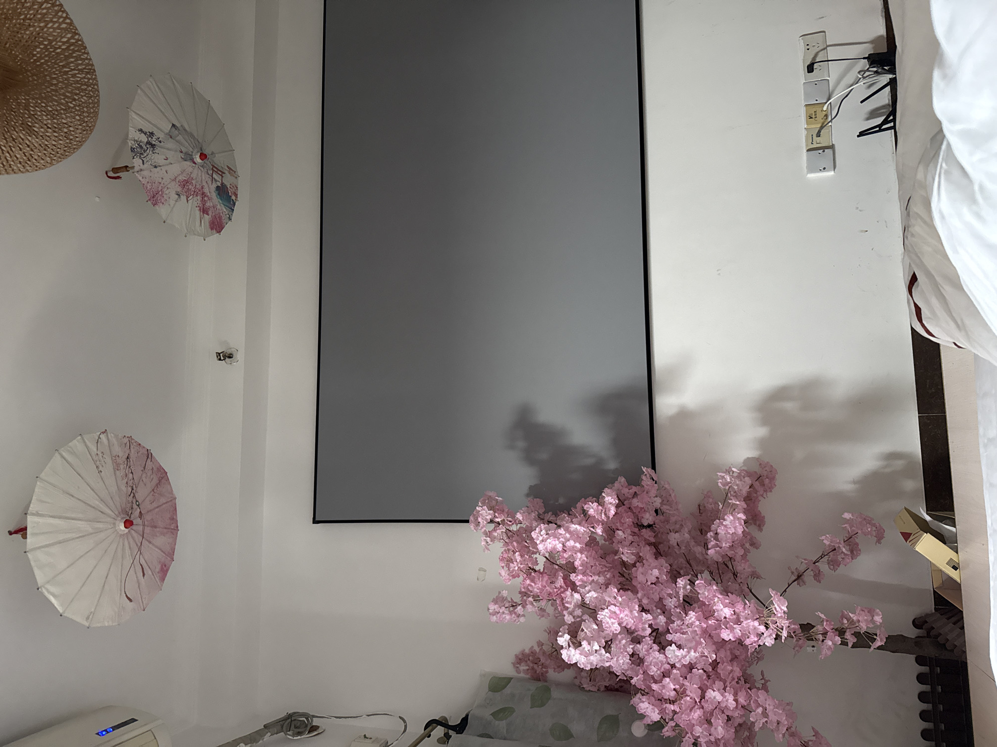 A dimly lit room features a large, dark gray projection screen mounted on a white wall. To the left of the screen, a faux cherry blossom tree with vibrant pink flowers is positioned. Above the screen, on the ceiling, two decorative paper umbrellas are hanging, one with pink floral patterns and the other with colorful bird and floral designs. A woven straw hat is partially visible hanging from the ceiling on the right side. Near the floor, electrical outlets with various plugs are visible on the white wall. A white bed with white linens is in the foreground, partially obscuring the bottom of the scene. The room appears to be a bedroom or living area within a residential or hotel setting. The image likely captures a scene within Haizhu District, China, given the context.