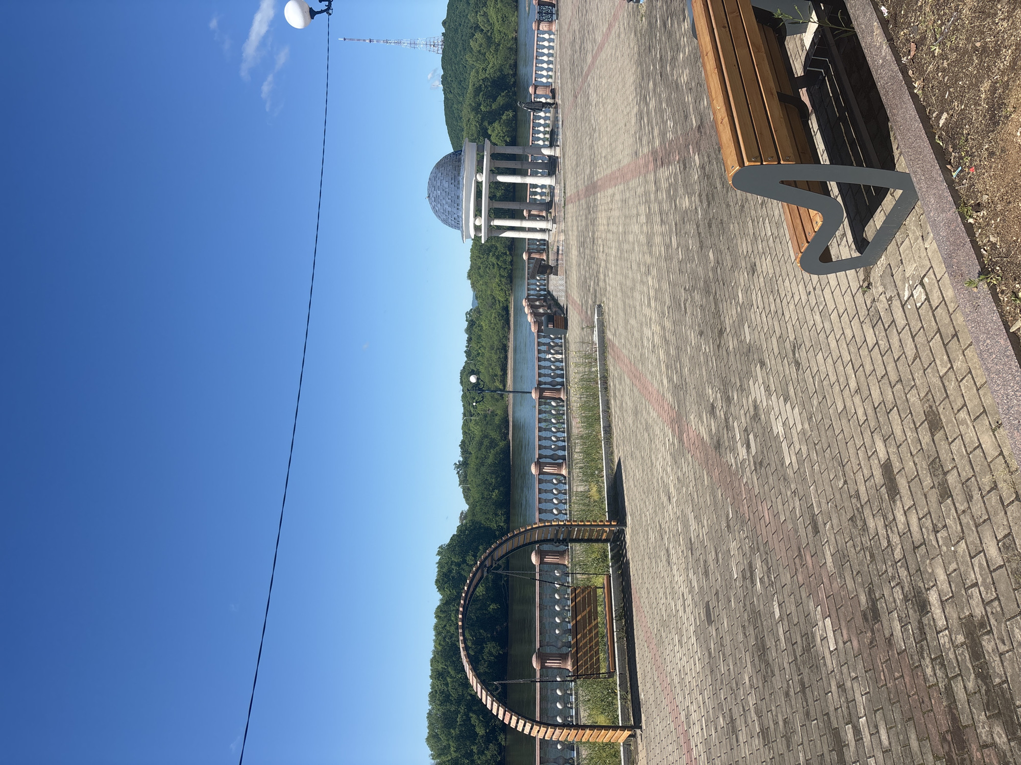 This image, captured in Birobidzhan, Russia, presents a peaceful and well-maintained waterfront promenade under a clear, bright blue sky. The scene is devoid of people, conveying a sense of tranquility.
The promenade is paved with interlocking bricks in shades of grey and reddish-brown. In the foreground on the right, a modern bench features wooden slats and a distinctive wavy grey metal support frame, casting a sharp shadow. Further along the walkway, a long, curved wooden bench with a metal frame is positioned to face the water. The edge of the promenade is lined with white, classical-style balustrades with dark detailing, overlooking the calm water.
A prominent architectural feature in the mid-ground is a white classical gazebo with a grey, domed roof, supported by columns. Beyond the promenade and the balustrades, a placid body of water, likely a river, reflects the sky. The opposite bank is covered with a dense, lush green forest that extends up a distant hill. A tall, slender metal lattice tower, possibly a communications tower, rises from the top of the forested hill.
The sky is a brilliant, cloudless blue, with only a few wisps of white clouds visible in some cropped sections. A spherical white street lamp on a black pole is visible in the upper left corner. The strong, defined shadows indicate either morning or early afternoon. There is no visible text in the image.