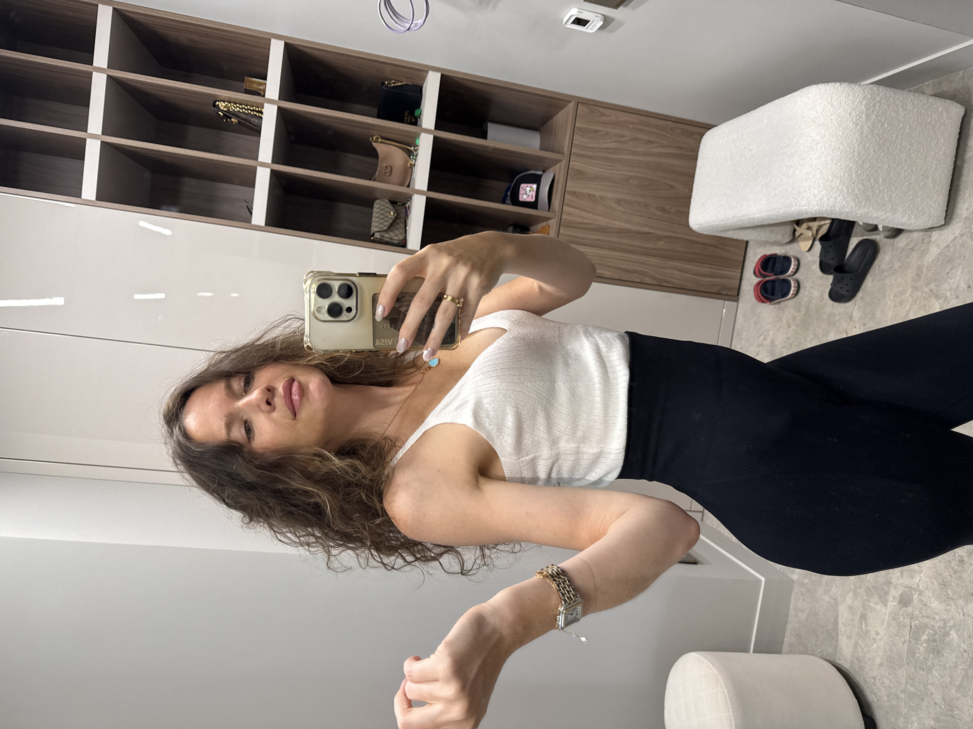 A woman is taking a mirror selfie in what appears to be a dressing room or closet. She is wearing a white ribbed tank top and black leggings. Her curly brown hair is down, and she is wearing jewelry, including a necklace with a pendant and rings. The background shows a large mirror, a closet with shelves displaying handbags and a hat, and a seating bench with shoes on the floor. To the left, there is a white upholstered stool. The floor is tiled. The image does not contain information to determine the location of Khimki, Russia.