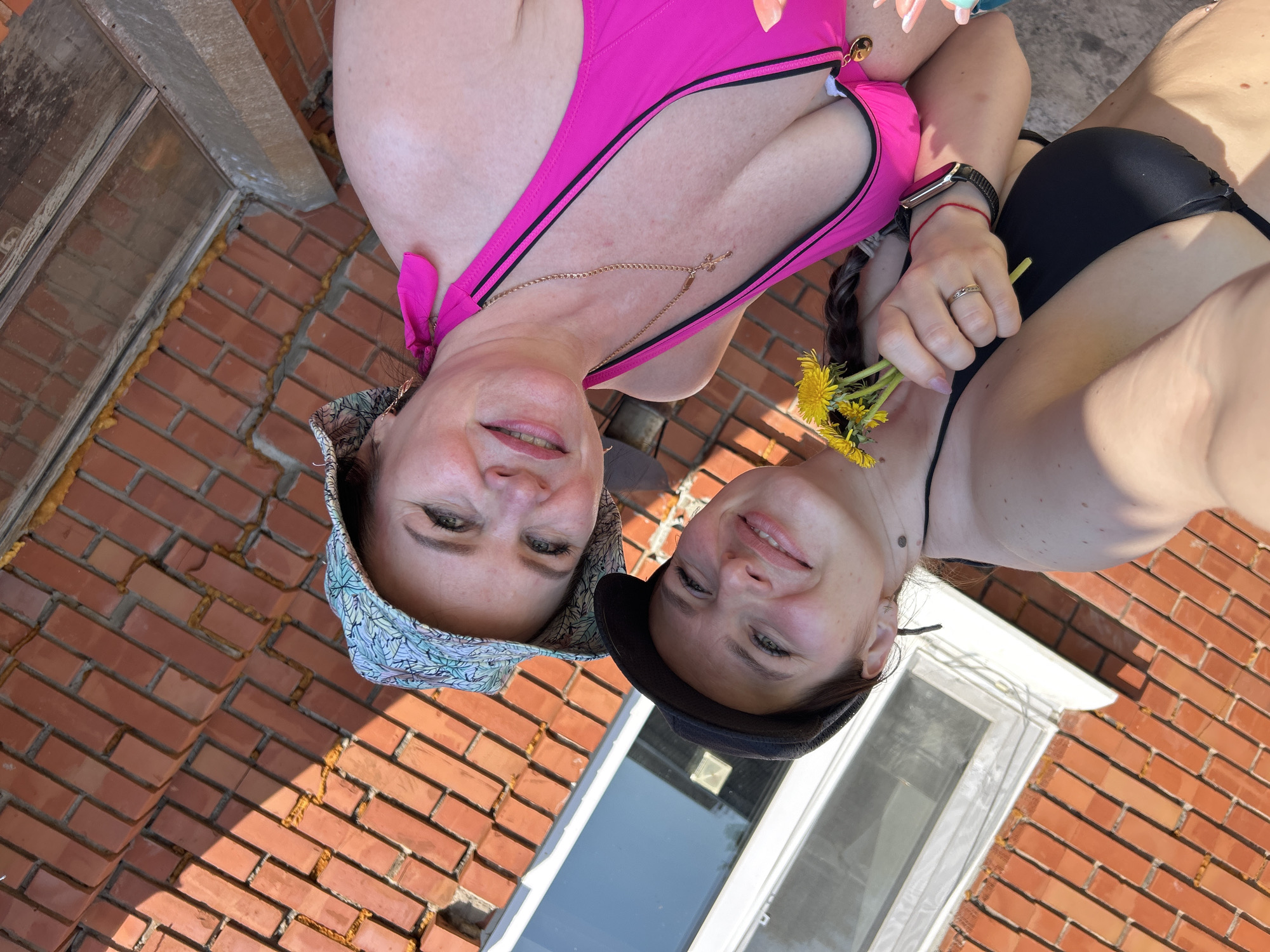 The image depicts two women posing for a selfie outdoors in bright sunlight. The woman on the left is wearing a floral-patterned hat and a pink swimsuit. She has a gold necklace. The woman on the right is wearing a black top and holding a small bouquet of yellow dandelions. She is wearing a smartwatch on her left wrist and a ring on her finger. The background shows a red brick surface, likely a wall or ground, and a window with white trim. The location is presumed to be Rusalevka, Russia. The women are smiling and appear to be engaged in a recreational activity.