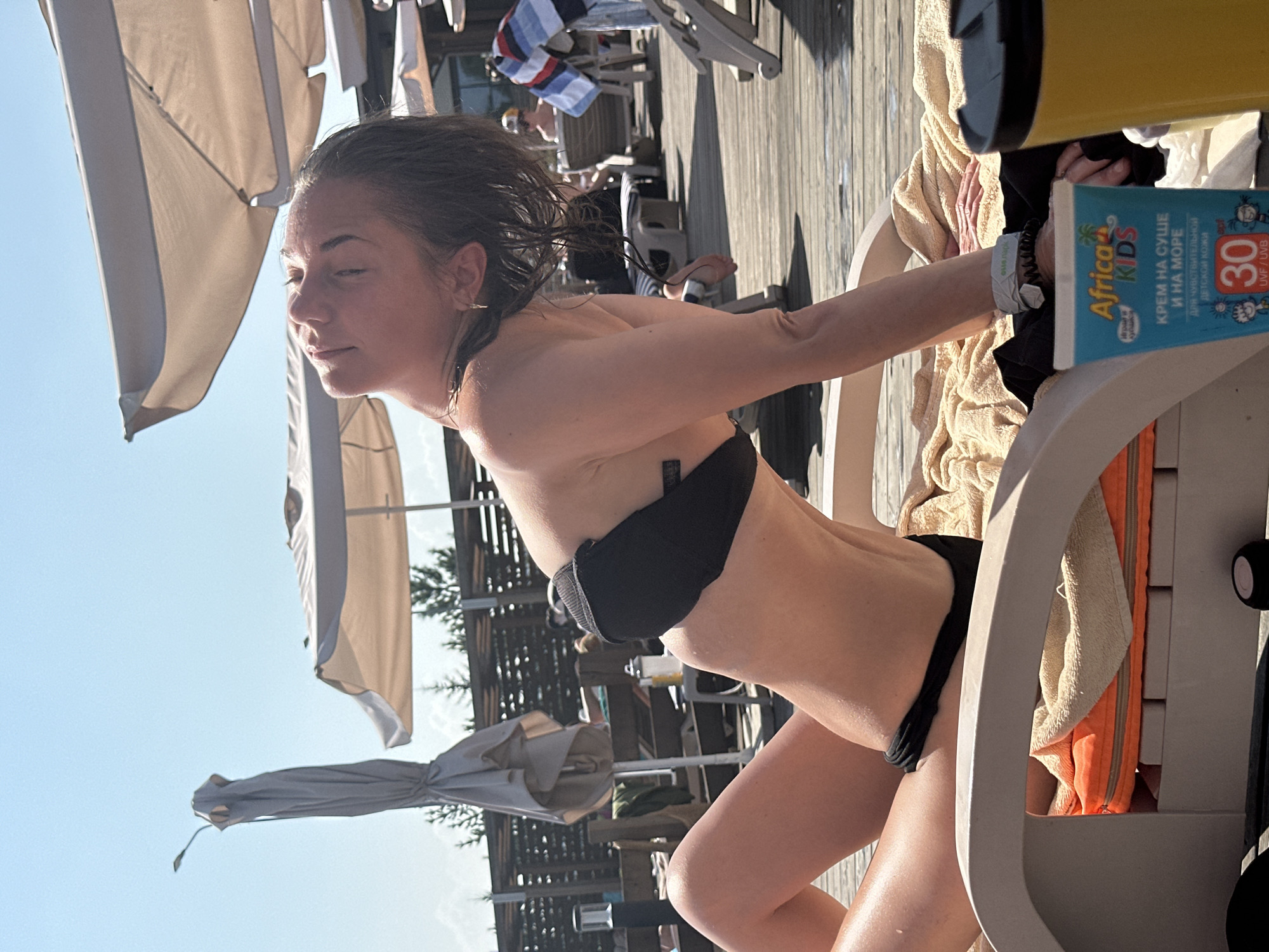 A young woman with wet hair, wearing a black bikini, is sunbathing on a lounge chair. She is looking up at the sky with her eyes closed, enjoying the sun. The lounge chair is placed on a wooden deck, and there are other lounge chairs and umbrellas in the background, suggesting a pool or beach resort. A bottle of sunscreen and a yellow cup are visible on the lounge chair next to her. The lighting suggests it is daytime and the weather is sunny.