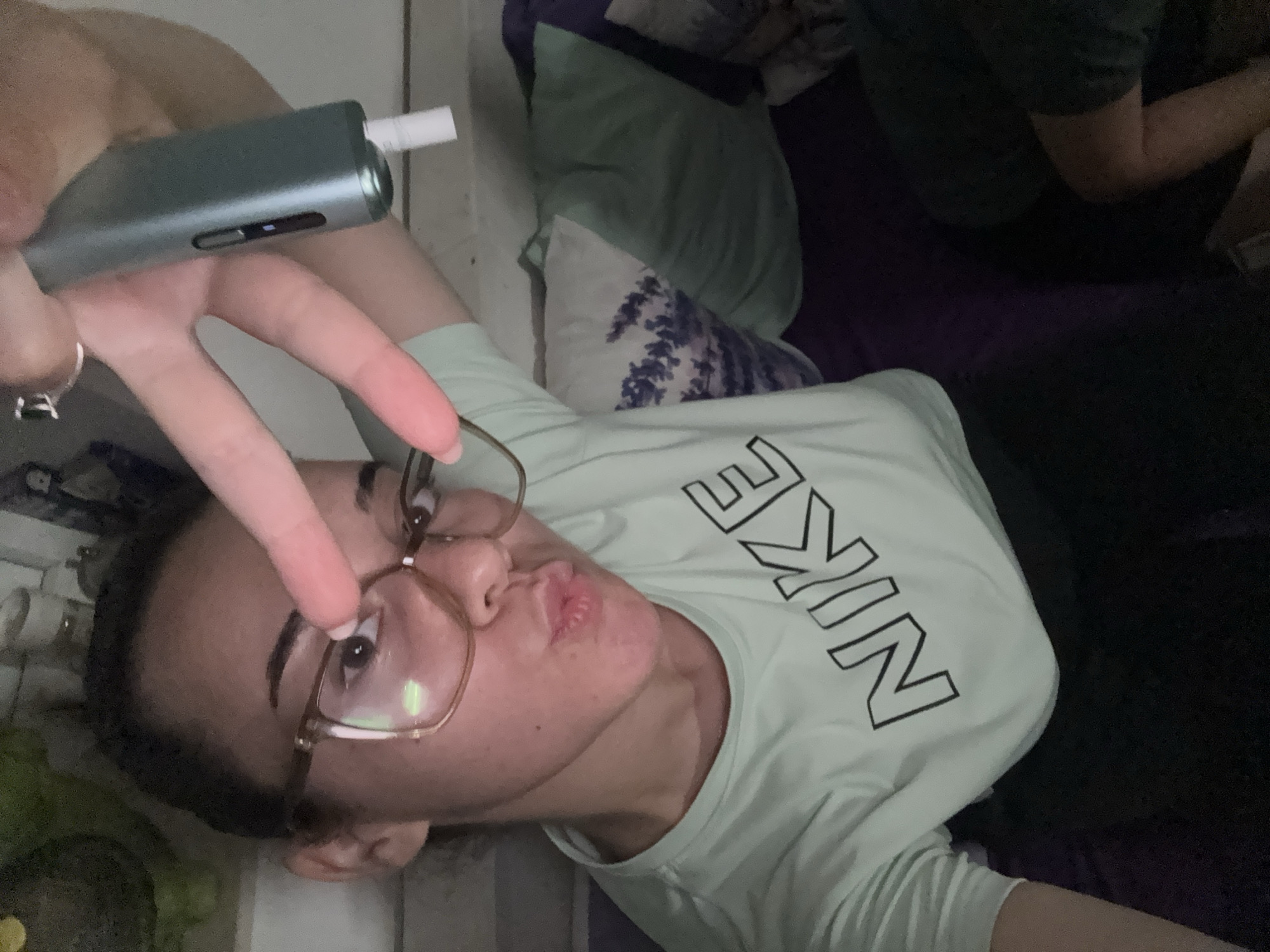The image shows a close-up, selfie-style shot of a young woman with dark hair pulled back. She is wearing large, gold-rimmed glasses and a light green t-shirt with the Nike logo in black. Her mouth is pursed, and she is holding up her right hand with her index and middle fingers extended in a peace sign. In her left hand, she is holding a teal-colored vaping device with a white filter. The background appears to be a dimly lit indoor setting, possibly a living room, with a purple couch and pillows visible. There are no discernible city or location details.