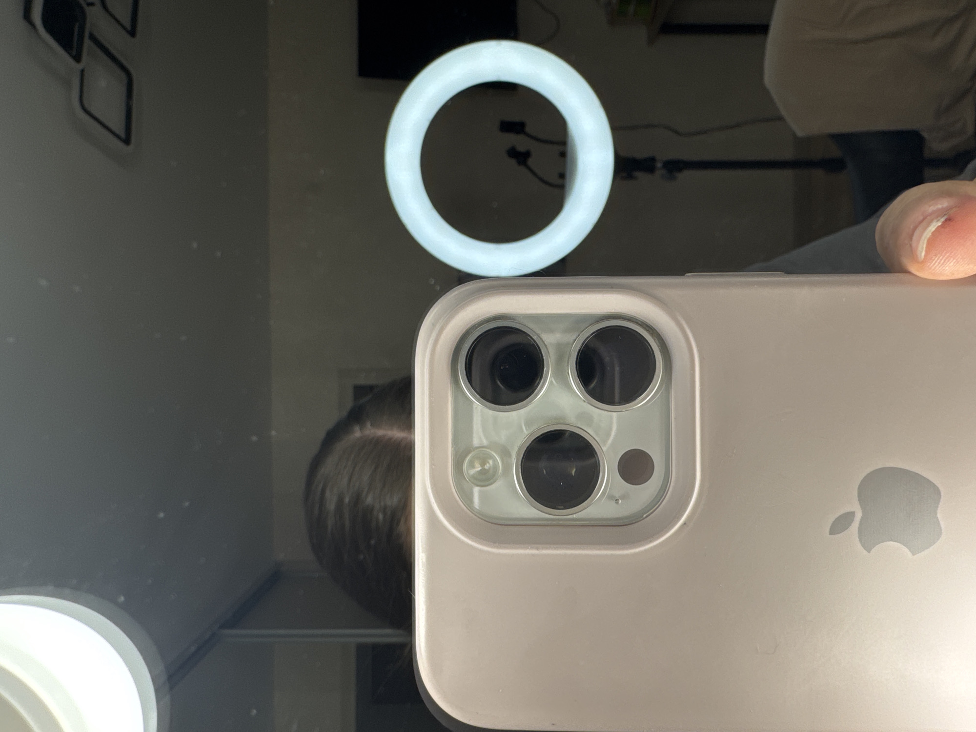 The image shows a close-up of a light pink smartphone with a triple-camera system. The phone is held in a hand, and the reflection in a mirror behind it shows a person's head and hair, along with a ring light. The background in the reflection is dimly lit and appears to be an indoor setting, possibly a room with furniture and a black television screen. There is no indication of the city of Saratov, Russia, or any specific geographic location in the image.