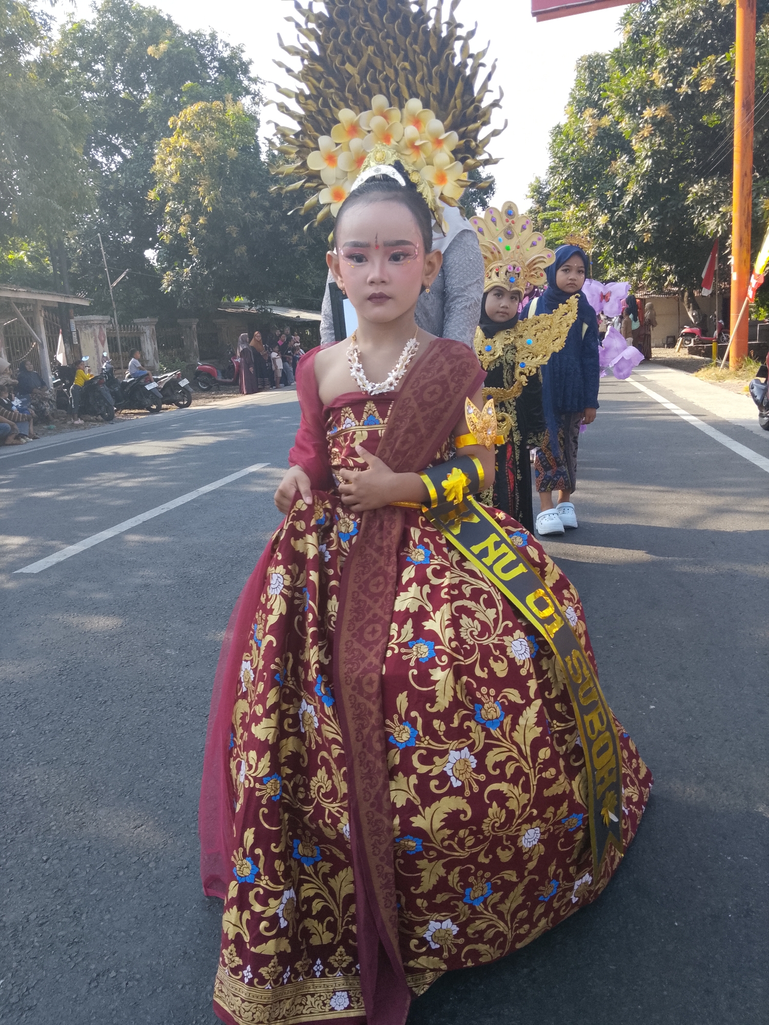 A young girl is dressed in a traditional costume, standing on a street in Buduan, Indonesia. She has elaborate makeup on her face, including painted eyebrows and eyeshadow. Her costume consists of a maroon dress with gold floral patterns and a sheer red overlay. She also wears a white beaded necklace and a sash across her torso that reads "NUO SUBO R" in gold letters. She has a golden crown-like headpiece adorned with white and yellow flowers, and a large headdress that extends upwards.
Behind her, two other children are also in traditional attire. The child immediately behind her wears a black outfit with a golden headpiece and a dark blue headscarf. The child further back is wearing a dark blue headscarf and a dark blue jacket over a patterned outfit.
The street is paved with asphalt and has a white line marking the lane. In the background, there are trees, buildings, and parked motorcycles. Several people are visible on the side of the street, watching the parade or event. The sun is shining, casting shadows on the road. The scene appears to be part of a cultural procession or festival.