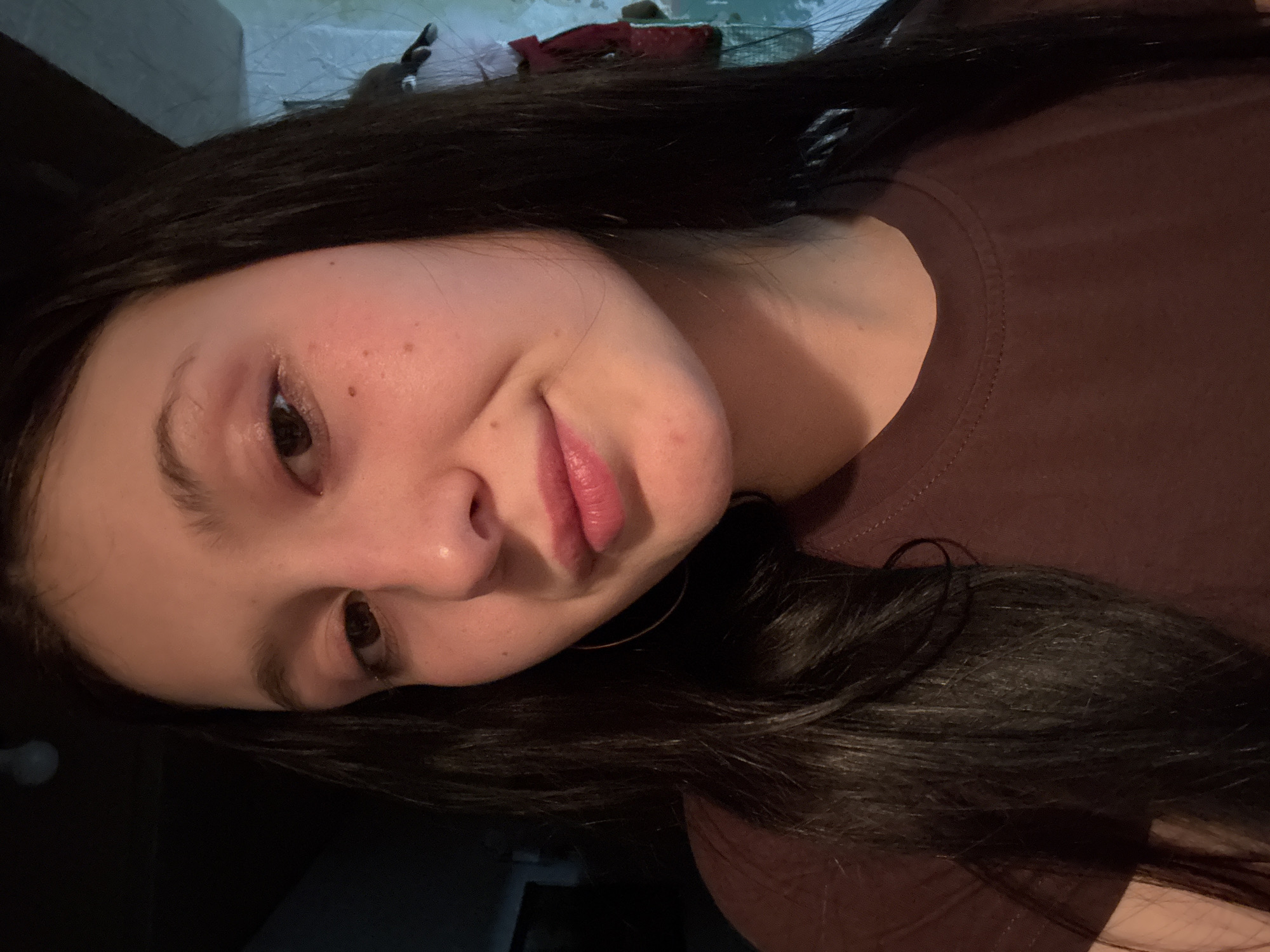 A young person, likely female, with long dark hair, is captured in a close-up, slightly tilted portrait, possibly a selfie. She is looking directly towards the viewer with dark eyes and a soft, slight smile, her lips a rosy pink. Her complexion shows some freckles and natural variations. She appears to be wearing a dark reddish-brown, crew-neck t-shirt. A golden hoop earring is visible on her right ear (viewer's left). The lighting is soft and appears to be artificial or low light, casting subtle shadows and highlighting her features. The background is indistinct, showing muted tones of light blue, grey, and dark brown, suggesting an indoor setting with minimal distractions. No specific activities are depicted other than posing for the photo. There is no visible text. The location context provided is Фоки, Russia.