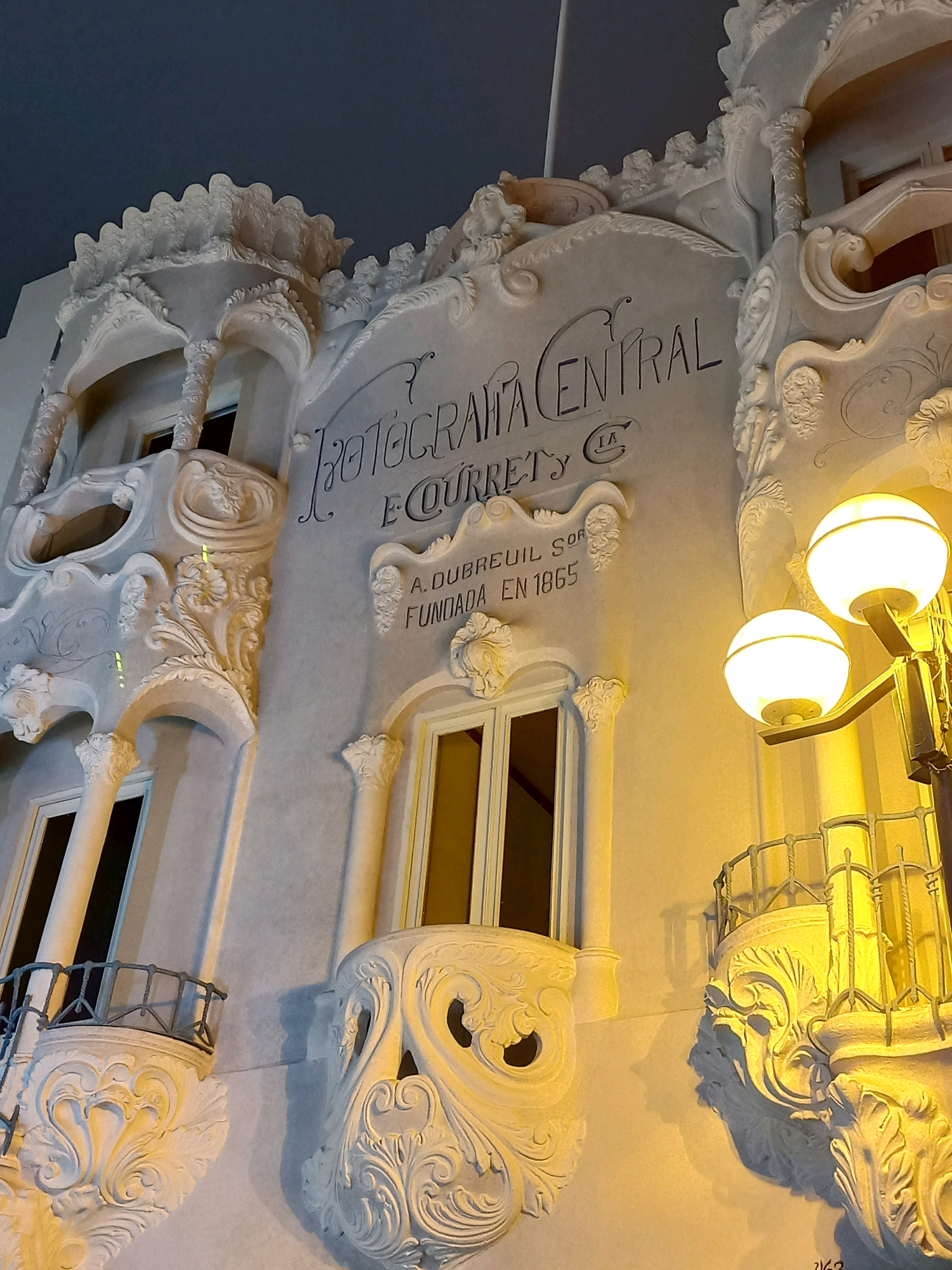 The image shows the exterior of a building with ornate, white stucco decorations and text carved into the facade. The text reads "FOTOGRAFIA CENTRAL E. COURRET Y C. A. DUBREUIL SOR FUNDADA EN 1865". The building features decorative balconies with intricate scrollwork and wrought-iron railings. A street lamp with two globes illuminates the scene with warm, yellow light. The architecture appears to be historic and possibly art nouveau in style. The overall impression is one of a well-preserved historical facade.
This building is located in the historic center of Lima, Peru, specifically at Jirón de la Unión 586. It is known as the "Casa Courret," which was once the studio of the renowned French photographer Lucas Courret and later housed his family's photographic business. The inscription refers to the establishment of the Courret photography business by A. Dubreuil in 1865.