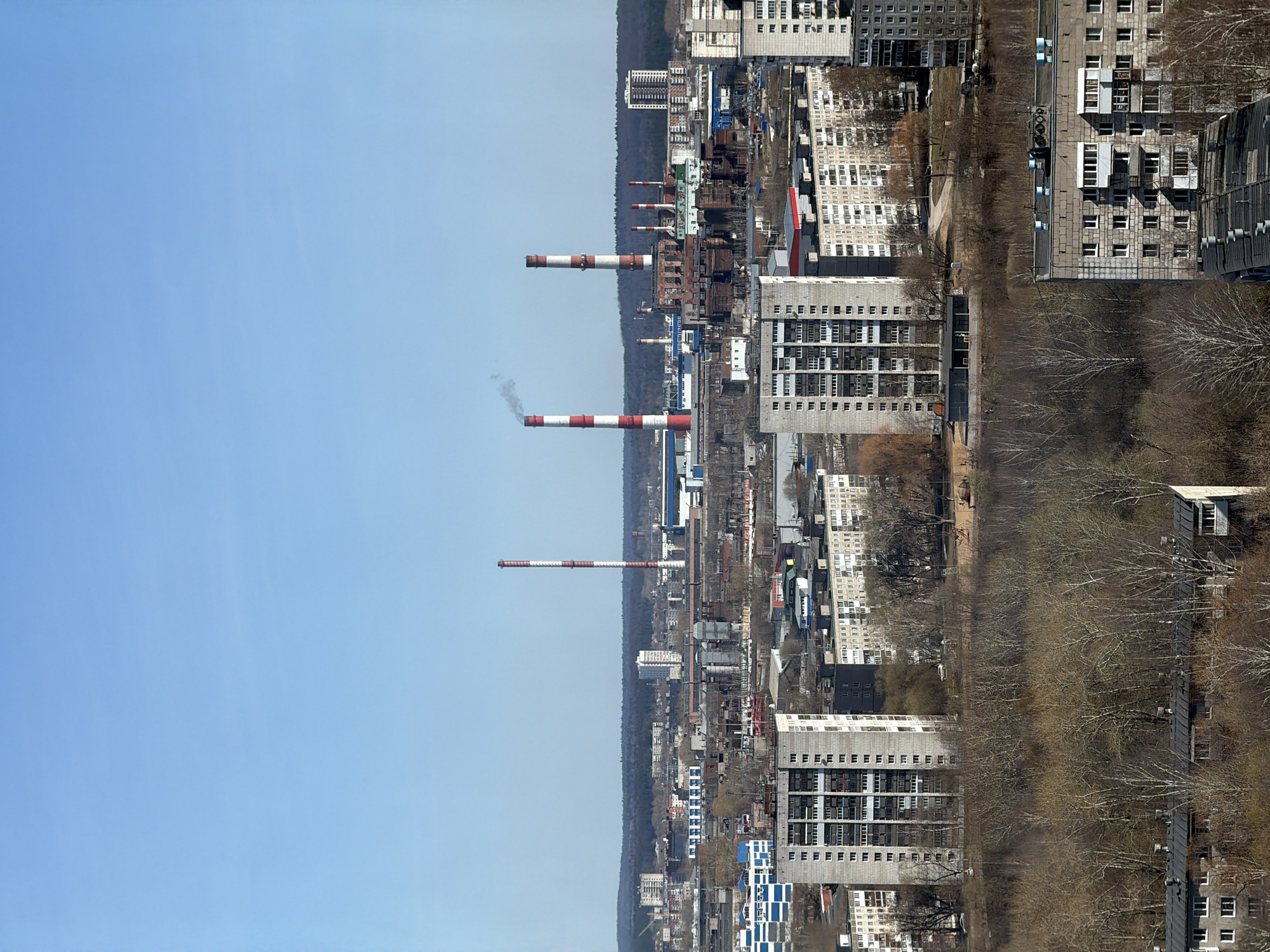 This image captures an urban scene in Perm, Russia, featuring several industrial chimneys with red and white stripes extending into a clear blue sky. One of the chimneys is emitting smoke. Below, the cityscape reveals a mix of high-rise residential buildings and industrial structures. A band of trees, mostly bare, runs along the right edge of the image, suggesting a park or green space adjacent to the built environment. The layout suggests a dense urban area with industrial activity.