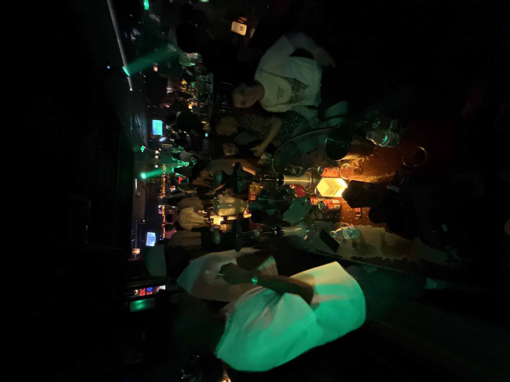 The image shows a dimly lit interior, likely a bar or nightclub, with green and blue lighting. Several people are visible, some standing and some seated at tables. In the foreground, a person in a white shirt is partially visible, illuminated by green light. A long table is set up with various items, including bottles, glasses, and shisha pipes. The atmosphere appears to be social and lively, with people engaged in conversation and enjoying themselves. The overall scene depicts a night out in a vibrant urban setting.