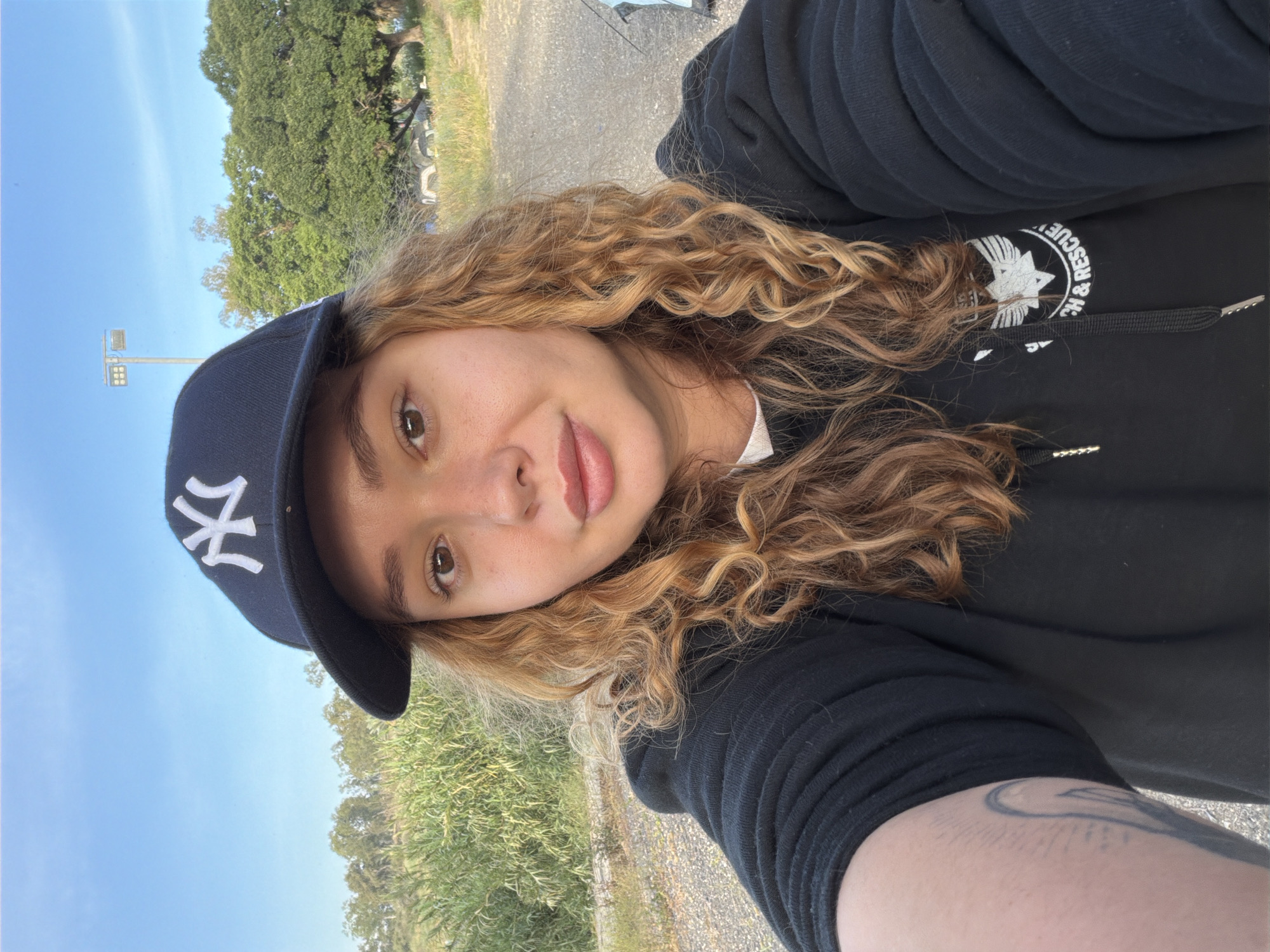 The image is a selfie of a young woman wearing a black New York Yankees baseball cap and a black hooded sweatshirt with the words "H & Rescue" printed on it. She has curly, light brown hair and appears to be outdoors. The background shows a blue sky, trees, and some vegetation. The location is in Emek HaYarden Regional Council, Israel.