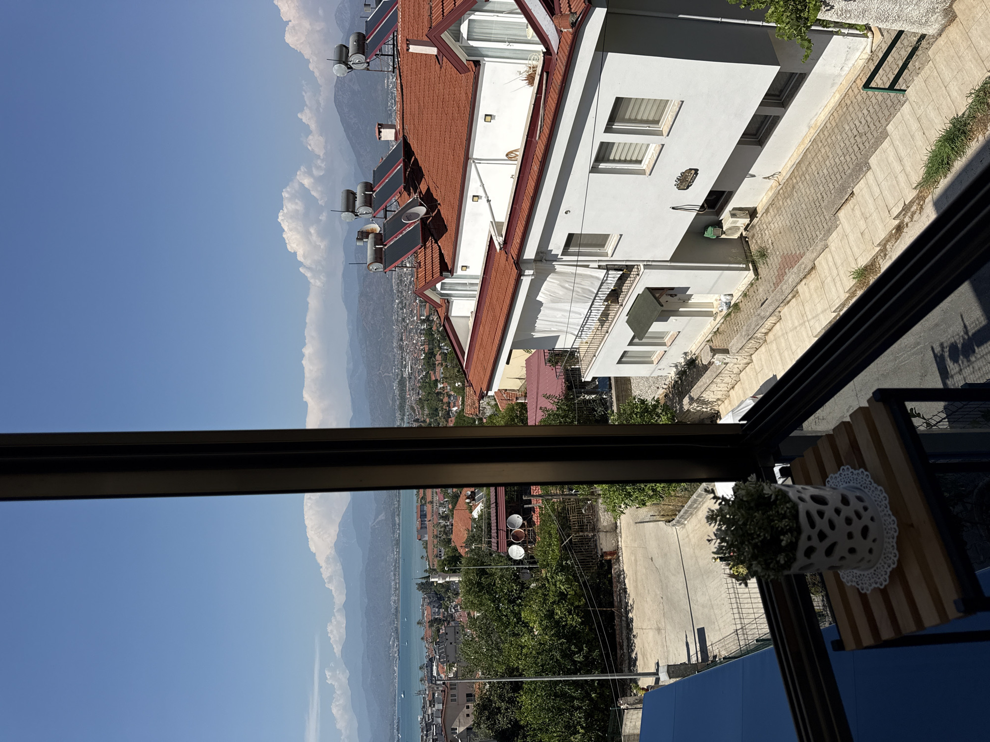 This is a tilted, medium-angle shot taken from an elevated position, likely a balcony or window, looking out over a town nestled against a body of water and mountains. The scene depicts a bright, sunny day with a clear blue sky and scattered white clouds.
In the foreground, a dark frame, possibly a window or railing, bisects the image. To the right of this frame, a wooden table with a decorative white planter containing a small plant is visible.
The midground and background reveal a town with red-tiled roofs and white buildings. Several solar water heaters are visible on the roof of one building. A body of water, likely a sea or large lake, stretches towards a distant mountain range. The mountains are hazy, suggesting a warm, clear day. The town appears to be built on a slope leading down to the water.
The overall impression is one of a peaceful, sunny day in a coastal town. The presence of solar water heaters suggests a focus on renewable energy. The scene evokes a sense of warmth and relaxation. No people are visible in the image, and there is no discernible text.