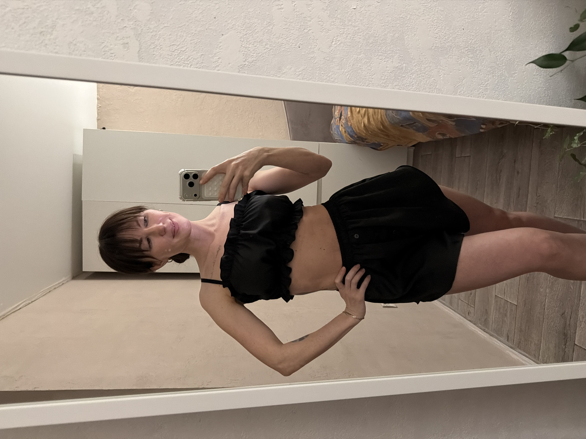 A woman is reflected in a mirror, holding a phone to take a selfie. She is wearing a black, ruffled top and black shorts. The background includes a white cabinet, a wall, wooden flooring, and a plant. The location is Perm, Russia.