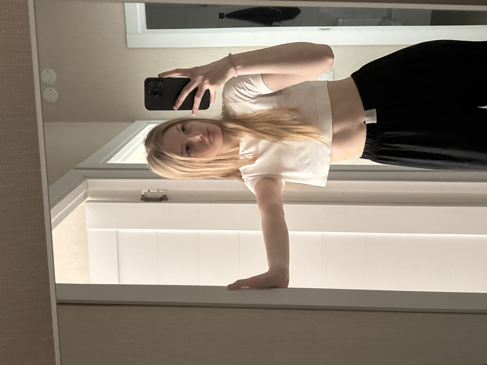 A young woman with blonde hair is taking a mirror selfie in a room. She is wearing a white crop top and black wide-leg pants. She is leaning against a doorframe with her right arm. The room appears to be a bathroom or dressing area, with a mirror, a door, and a towel hanging on a hook in the background. The lighting is soft and natural. The image does not contain any specific details to indicate it is located in the city of Perm, Russia.