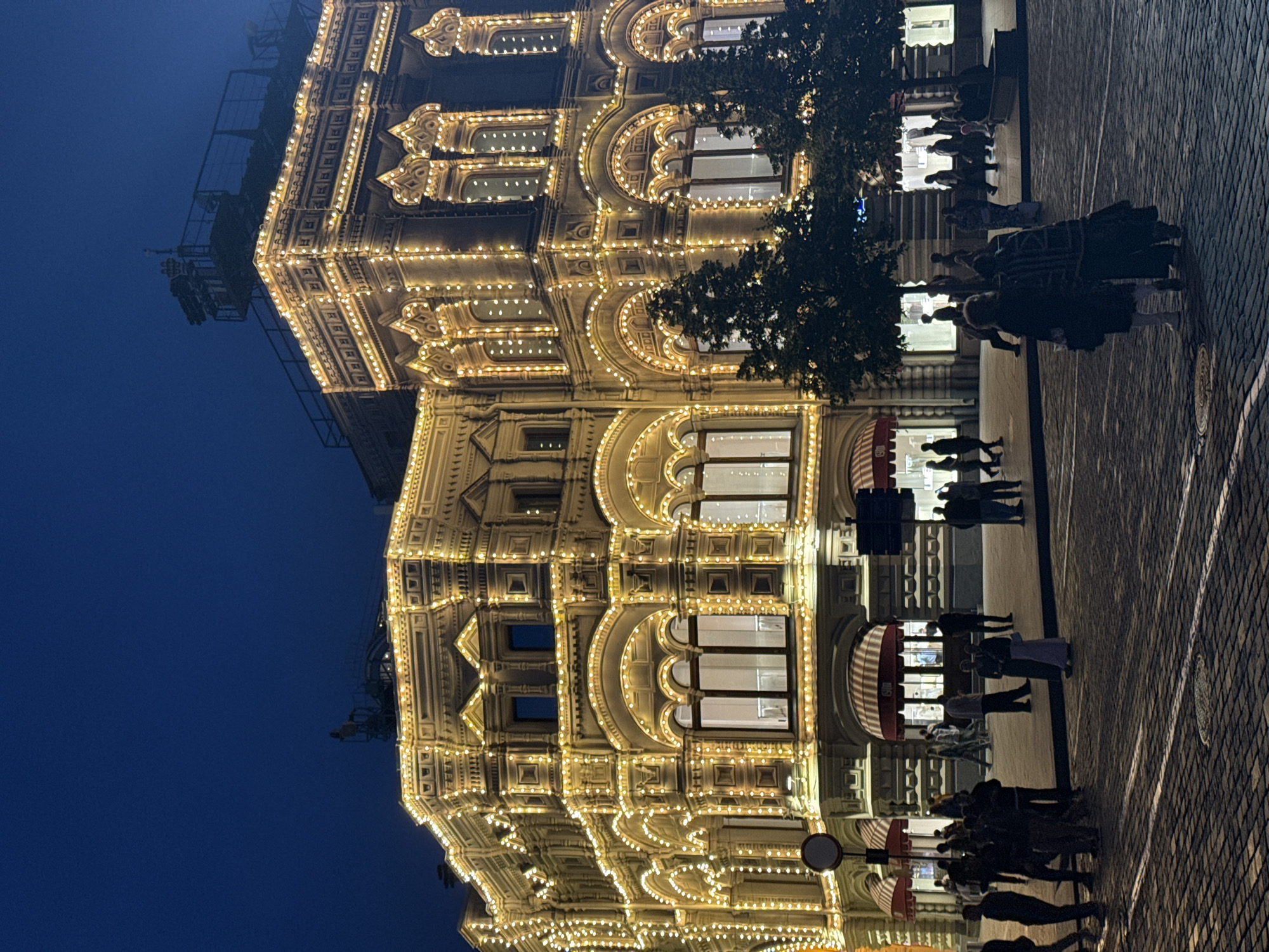 In an urban setting at night, a grand, ornate building is illuminated by strings of warm, golden lights that outline its architectural features. The building, with multiple stories, features arched windows and intricate decorative elements. In front of the building, a paved plaza is visible, cobblestone patterned. Several trees are present in front of the building, partially obscuring the lower levels. A group of people are scattered across the plaza, some walking, others standing. The sky above is a deep, dark blue.