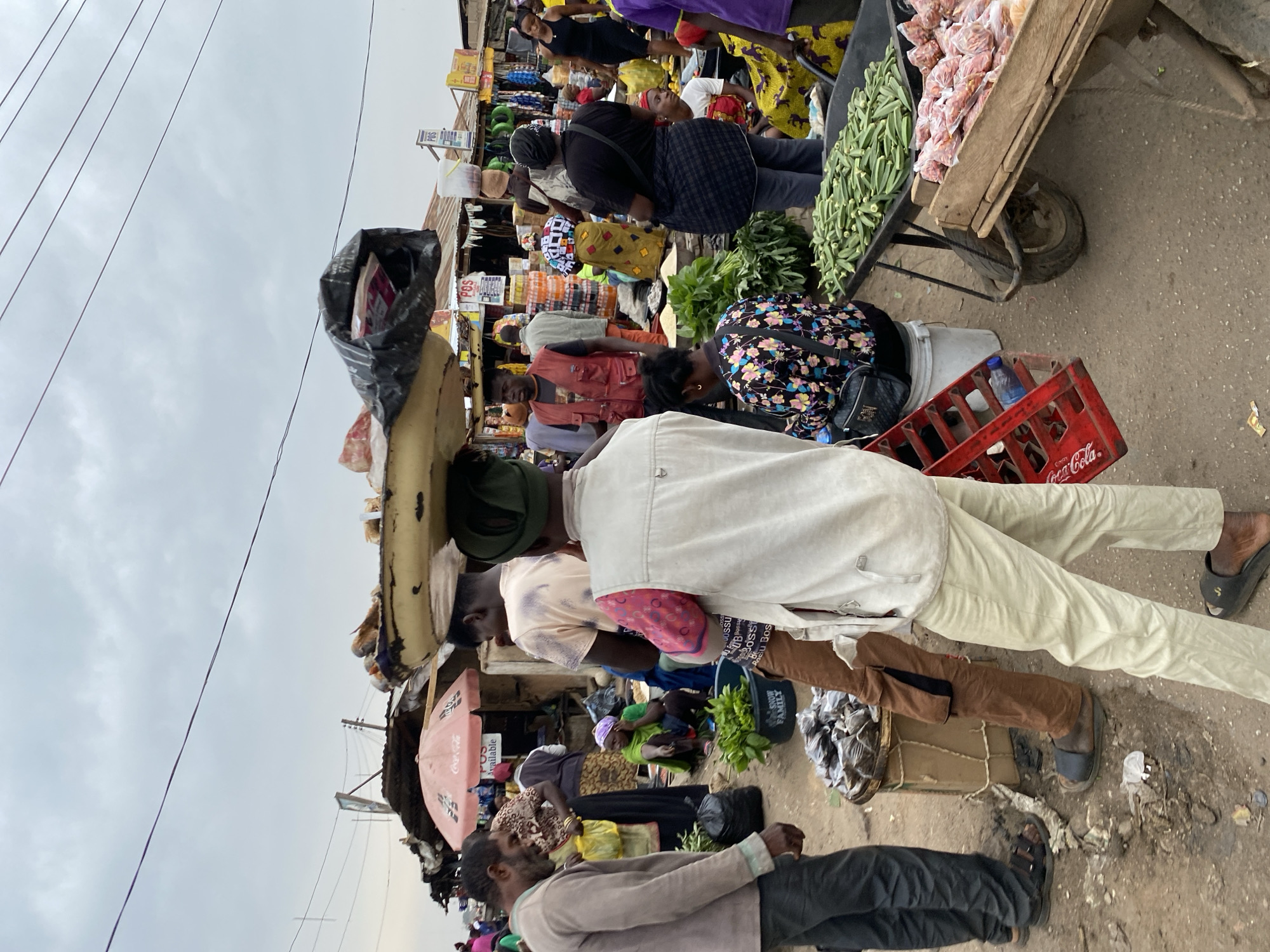 This image depicts a bustling market scene in Bukuru, Nigeria. The environment appears crowded and somewhat chaotic. Numerous people are present, engaging in various activities related to buying and selling goods. Some individuals are walking, while others are standing or squatting to examine merchandise.
Several vendors are displaying their wares, including fresh produce such as vegetables and bagged items. One vendor is transporting goods in a wheelbarrow. A man carries a load of goods balanced on his head, wrapped in black plastic. A red Coca-Cola crate lies on the ground. The sky is overcast.