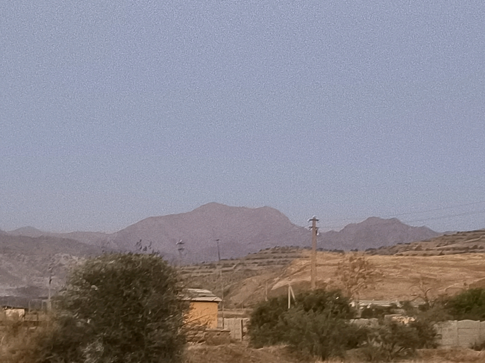 The image depicts a hazy, arid landscape with rolling hills and mountains in the background. The sky is a pale, washed-out blue. In the middle ground, there is a small, light orange building with a darker roof, partially obscured by trees. A wooden utility pole with wires is visible on the right side of the frame, and terraced hillsides suggest agricultural activity. A large, dense green tree is prominent in the lower left quadrant, and other scattered vegetation dots the terrain. The overall impression is one of a rural, undeveloped area, potentially in the vicinity of Namdanak, Uzbekistan, characterized by dry vegetation and a muted color palette.