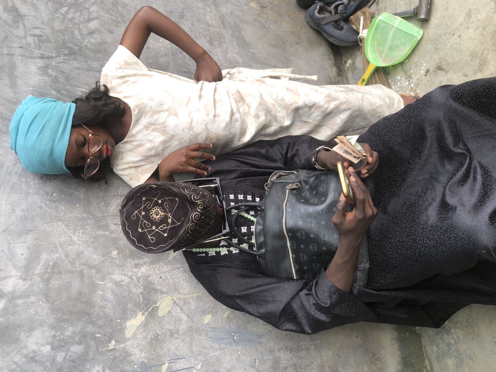 In Ado, Nigeria, a man wearing a black traditional outfit and a decorated cap is seated, looking at his phone and holding cash. A young girl, dressed in a cream-colored dress and a blue headscarf, stands beside him, leaning against a grey wall. She wears glasses and has orange lipstick on. The background features a textured grey wall and a concrete floor with some scattered white debris. A green dustpan, a broom, and a hammer are visible on the floor. A black handbag rests on the man's lap.