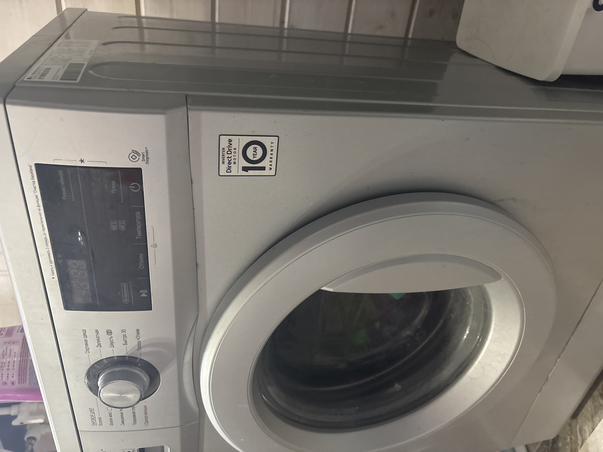 The image shows a close-up, slightly angled view of a silver LG washing machine. The machine is located in what appears to be a bathroom or laundry room, with tiled walls visible in the background. The control panel of the washing machine is visible, featuring a digital display, a rotary dial with various wash cycle options labeled in Russian (e.g., "Хлопок" - Cotton, "Спортивная одежда" - Sportswear, "Деликатная" - Delicates, "Шерсть" - Wool, "Быстро 30" - Quick 30), and buttons for power and other settings. A "10 YEAR WARRANTY" sticker with "INVERTER DIRECT DRIVE MOTOR" is prominently displayed on the right side of the machine. The washing machine door is slightly ajar, revealing the drum inside. The scene is lit by artificial light, and the overall impression is one of a functional appliance in a domestic setting. No specific location in Березка Снт (поварово), Russia can be identified from the image itself.