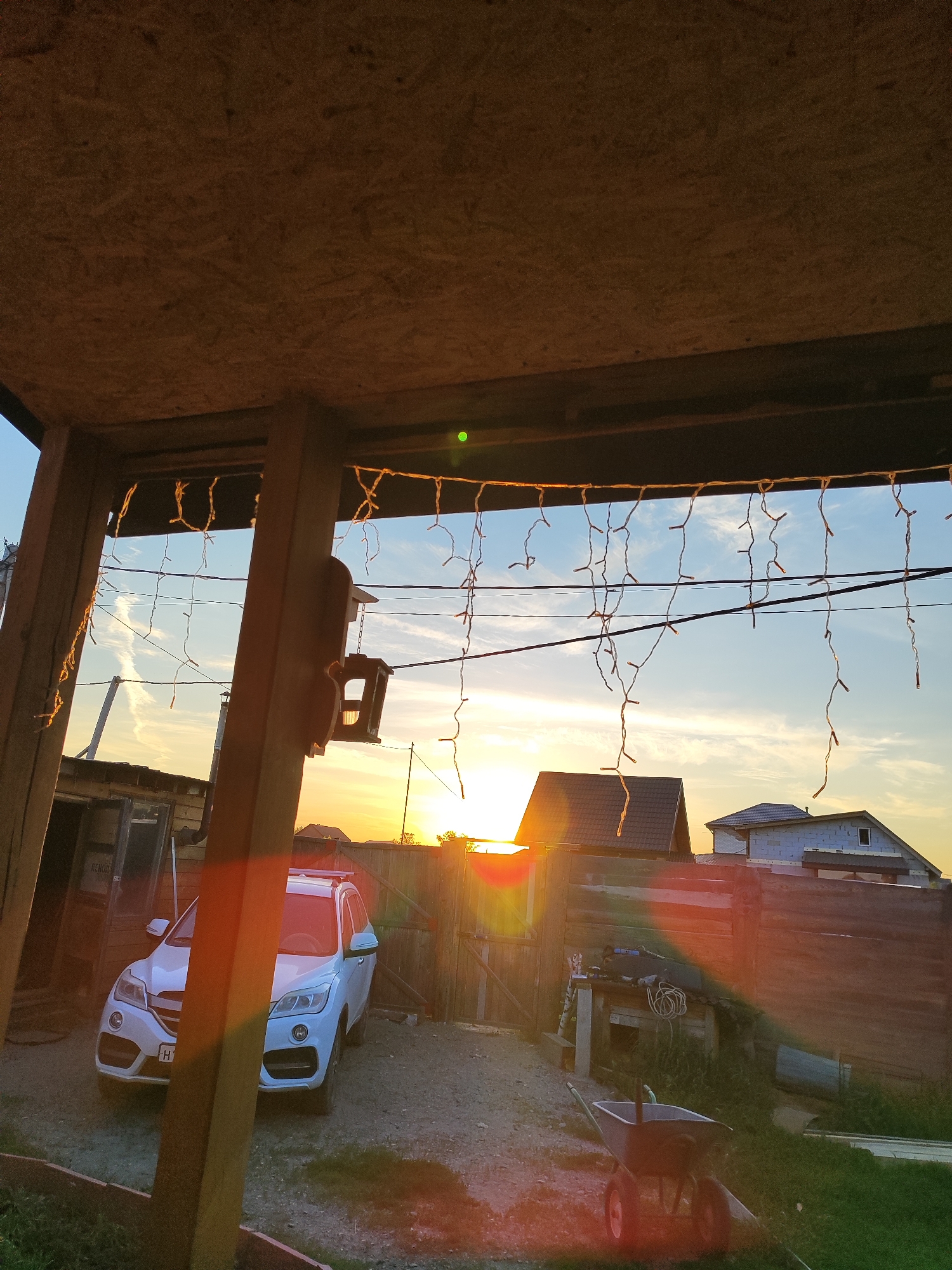 The image depicts a rural scene at sunset in Marusino, Russia. The foreground features a wooden structure with an OSB ceiling and supports, from which strings of decorative lights hang. A white SUV is parked in a dirt and gravel yard. Behind the car, a wooden fence separates the yard from other properties.
In the background, several houses are visible, some under construction. The sky is a clear blue with wispy clouds, and the setting sun casts a warm, golden glow across the landscape, creating lens flares and highlighting the edges of objects. A birdhouse is hanging from one of the wooden supports in the foreground. A wheelbarrow with two red wheels is also present in the yard, suggesting outdoor work or maintenance. The overall mood is peaceful and serene, characteristic of a rural evening. There is no visible text in the image.