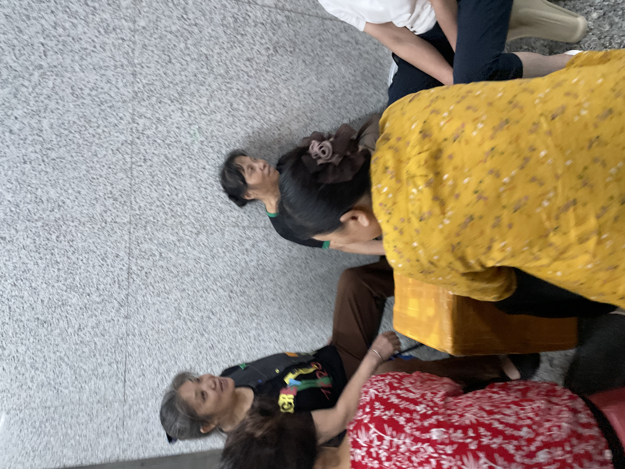 The image shows a group of people sitting on the floor or low stools against a textured gray wall. In the foreground, a person wearing a yellow floral print shirt is seen from behind. To their left, another person in a red and white patterned shirt is visible. Between them is a large, orange-wrapped rectangular object, possibly a package. Further back, a woman with graying hair tied back is seated, wearing a black t-shirt with a colorful graphic and a dark vest. Another woman in a black shirt is seated behind her, looking to the right. The environment appears to be indoors, possibly a hallway or waiting area. The location is identified as Liangjiang, China.