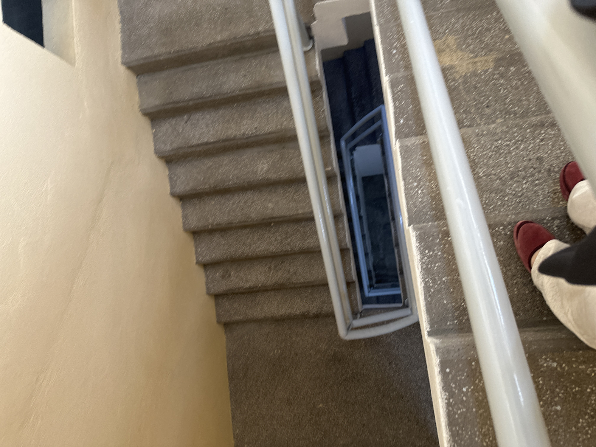 The image shows a view looking down a concrete staircase with a white handrail. The steps are made of speckled concrete, and the walls appear to be painted a light yellow. There is a rectangular opening in the center of the stairwell, revealing a dark interior. At the bottom of the image, a person's legs and feet are visible, wearing light-colored pants and dark red slippers. The person is standing on one of the steps. The scene is an indoor staircase, likely within a building. The image does not contain any specific visual cues to determine its location in Puebla, Mexico.