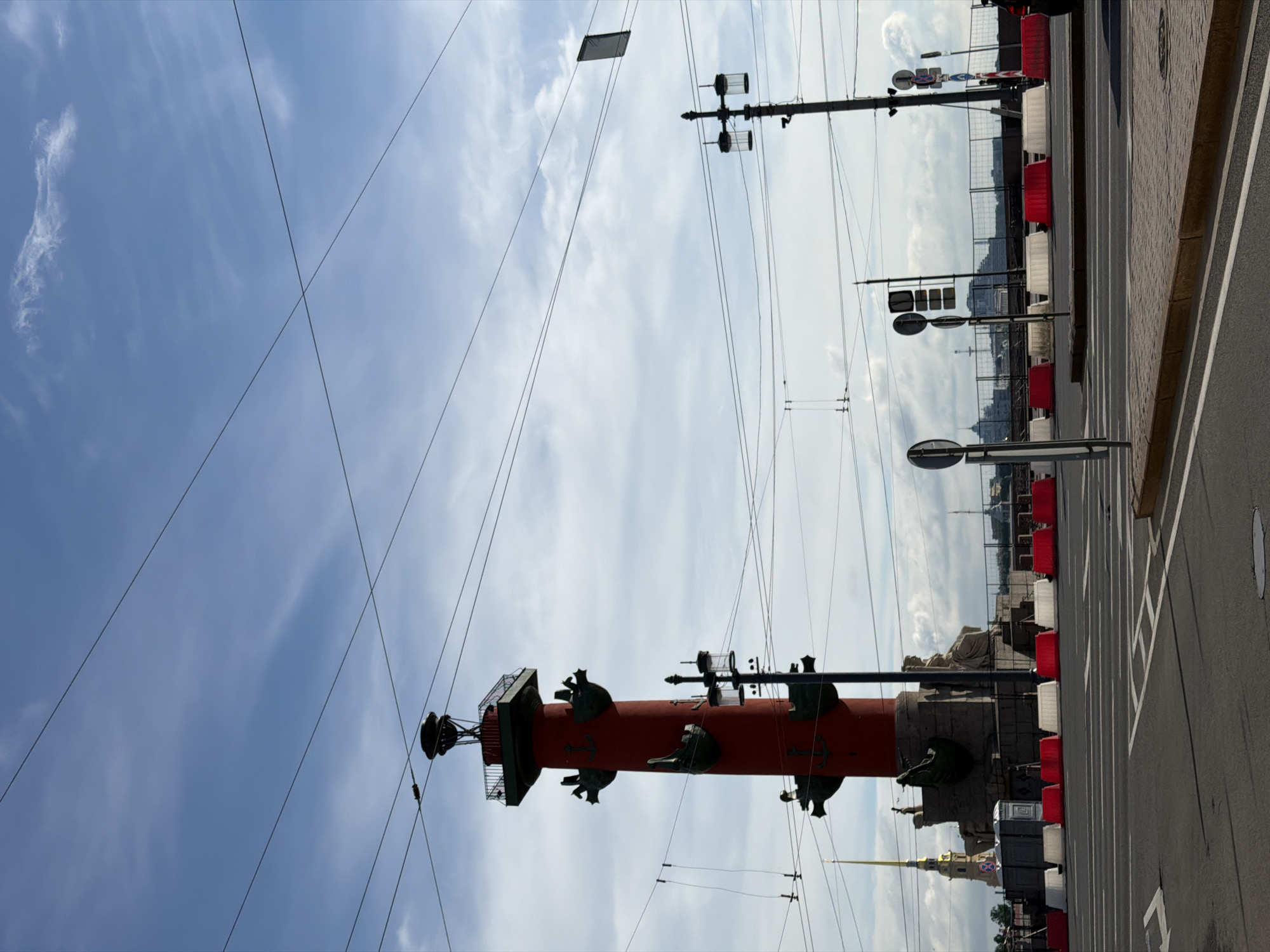 This is a medium shot, eye-level, exterior photograph of the Rostral Columns on the Spit of Vasilievsky Island in Saint Petersburg, Russia, on a cloudy day. The Rostral Columns are large, red, cylindrical structures topped with what appears to be beacons. They are decorated with anchors and ship prows. In front of the columns, a stone pedestal is visible, featuring several sculptures. To the left of the Rostral Columns, the spire of the Peter and Paul Fortress is visible in the background. The foreground shows a street intersection with road markings, concrete and red traffic barriers, and a sidewalk. Several streetlights and overhead wires are present throughout the scene. No people are visible in the photograph.