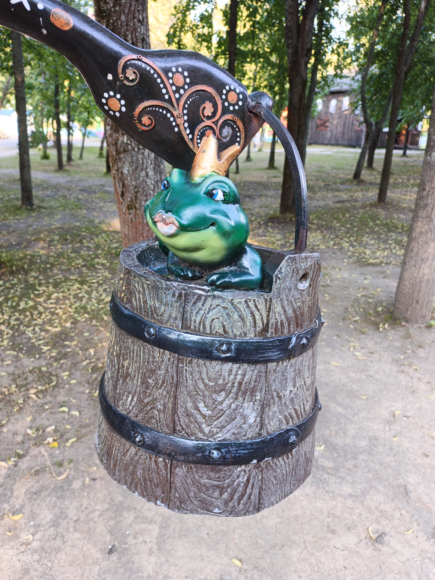 This is a close-up, medium shot of a decorative sculpture in a park-like setting. The sculpture features a green frog wearing a golden crown, peeking out of a wooden barrel. The frog has large, blue eyes and a slight smile. The barrel is dark brown with black metal bands. Above the barrel, a black, decorative element with gold and white embellishments hangs down, appearing to be part of a larger sculpture or structure, possibly a lever or handle, with a curved metal rod connecting it to the barrel. The background consists of trees with green foliage, casting shadows on the ground which is covered with fallen leaves and a concrete pathway. In the far background, a rustic wooden building is visible, suggesting a historical or themed park. The overall scene is outdoors and appears to be a sunny day. There are no people visible in the frame.
The sculpture of the frog and barrel is a well-known landmark in the city of Yaroslavl, Russia, located at the entrance to the Yaroslavl Zoo. It is a popular photo opportunity for visitors.