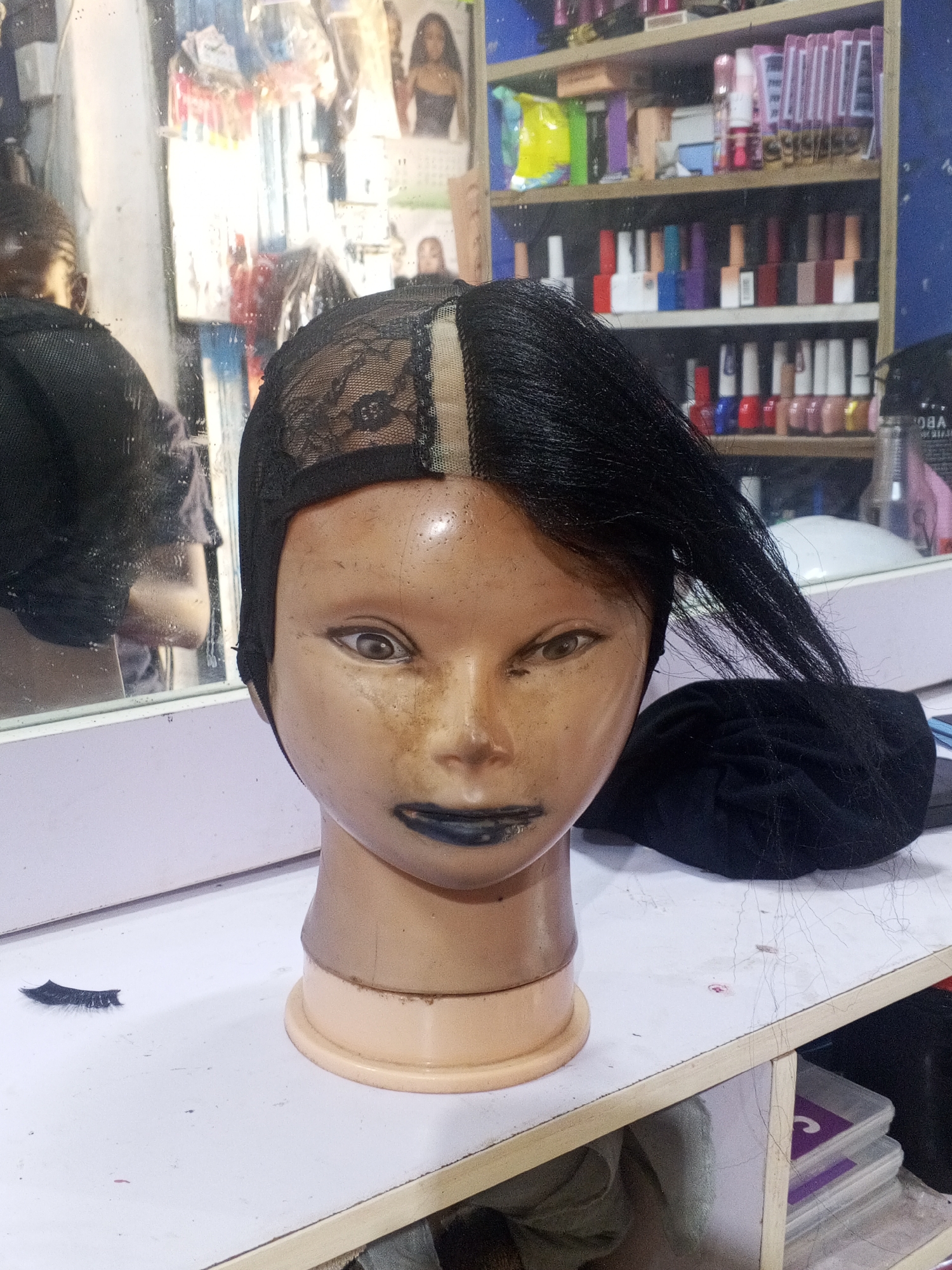 A mannequin head with a black lace wig cap and partially styled black hair rests on a white surface, likely a table, with a single false eyelash nearby. The mannequin has heavy black lipstick, and some discoloration on its face. In the background, there is a mirror reflecting a person wearing black, and a shelf stocked with nail polishes and other cosmetic items. The scene appears to be in a beauty salon or studio, suggested by the styling equipment and cosmetics present. The location is identified as Gwong, Nigeria, based on contextual information not explicitly visible in the image.