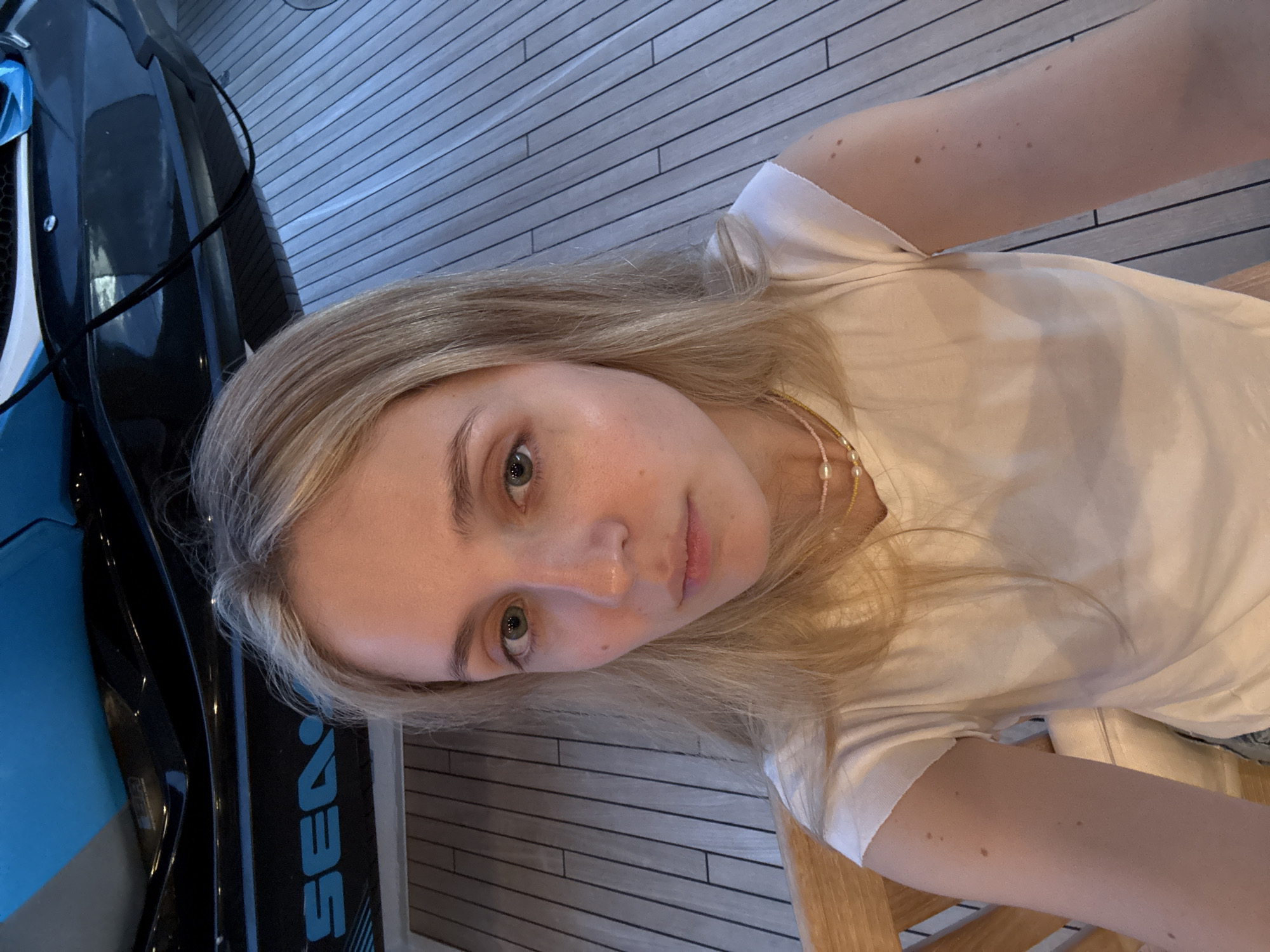 A light-skinned woman with blonde hair and light eyes is taking a selfie. She is wearing a white t-shirt and a beaded necklace. To her left is a blue and black Sea-Doo personal watercraft. The background appears to be a wooden deck with linear patterns. The location, Maravia, Mexico, cannot be determined from the visual information.