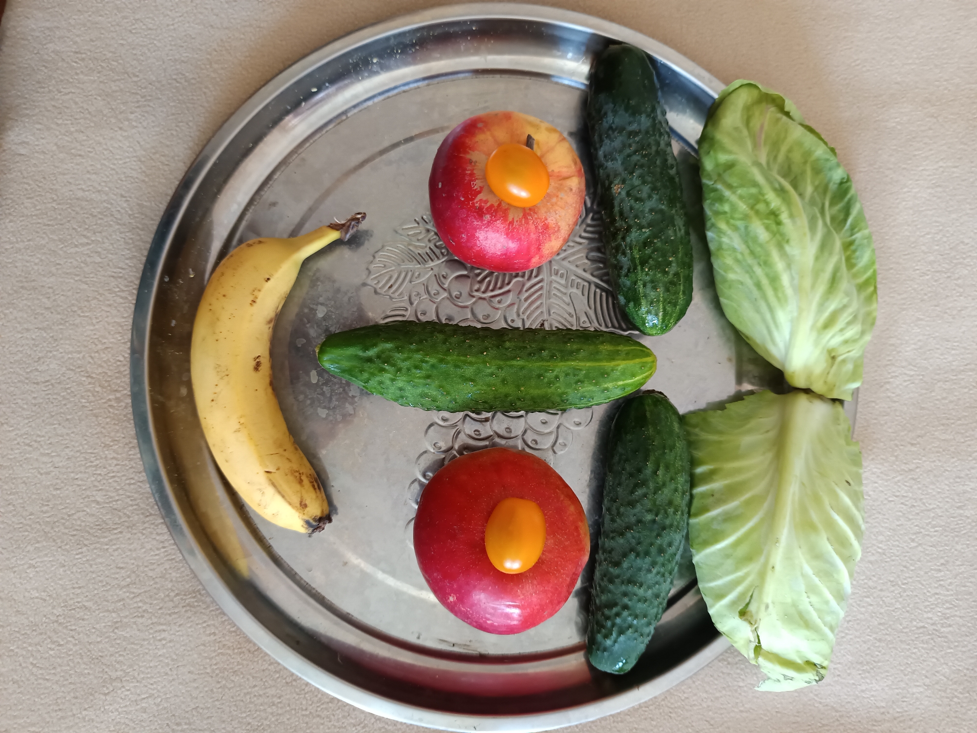 A still life image shows a variety of fruits and vegetables arranged on a silver platter. The platter is placed on a textured, light brown surface, possibly a tablecloth or countertop. The items on the platter include a yellow banana, two red apples each topped with a small yellow cherry tomato, three cucumbers, and two leaves of cabbage. The image is taken from a top-down perspective. The location is Tyshky, Ukraine.