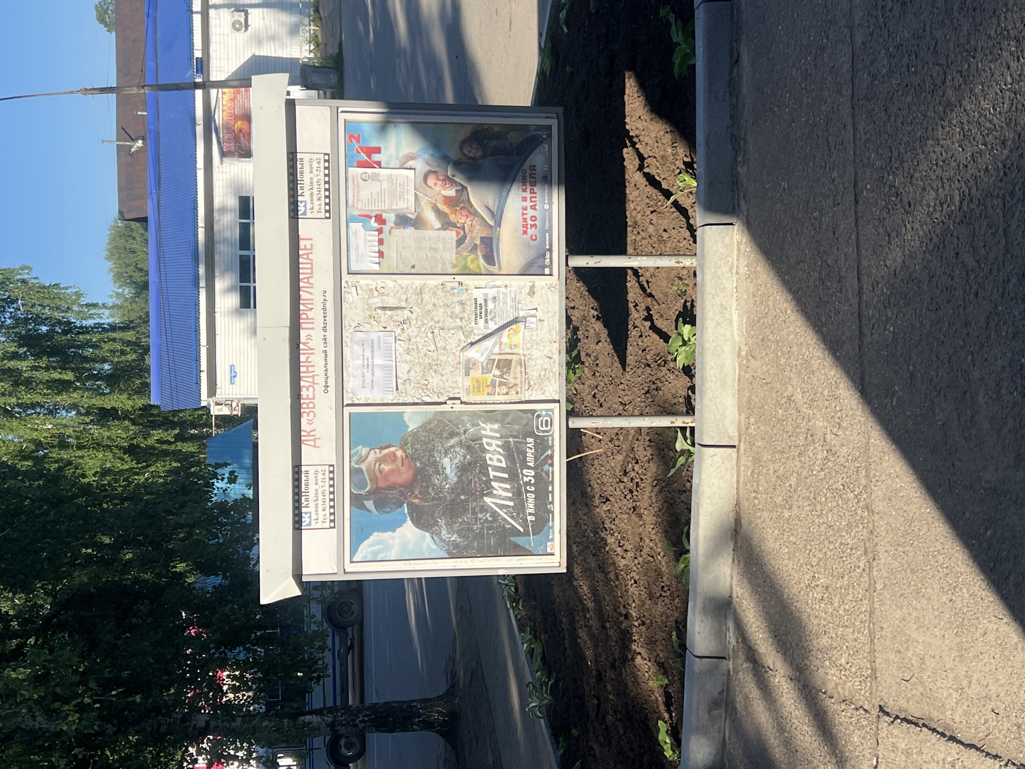 A freestanding movie poster display is positioned on a sidewalk in front of a building. The display has three sections showcasing different movie advertisements. The left section features a poster for the film "Литвяк" (Litvyak), depicting a woman in a pilot's helmet and goggles. The middle section has a collage of various smaller advertisements and notices. The right section displays a poster for a film with the title "Ан 2" (An 2), showing a group of people in a car. The top of the display has text in Russian that translates to "DK 'ZVEZDNIY' INVITES" and "Official website dkzvezdny.ru." Below this, the name of the cinema "КиноНовый" (KinoNoviy) is visible with contact information. The building behind the display appears to be a single-story structure with a blue corrugated metal roof and white siding. A sign for "ХОЗТОВАРЫ" (Household goods) is visible on the building. The scene is outdoors, under a clear blue sky with bright sunlight casting shadows. The display is surrounded by a flower bed with freshly turned soil and small green plants. The ground in front of the display is asphalt, with visible cracks and tire tracks. The context suggests this is a public area, likely in a town or city, given the presence of a building and sidewalk. The location specified is Noviy, Russia.