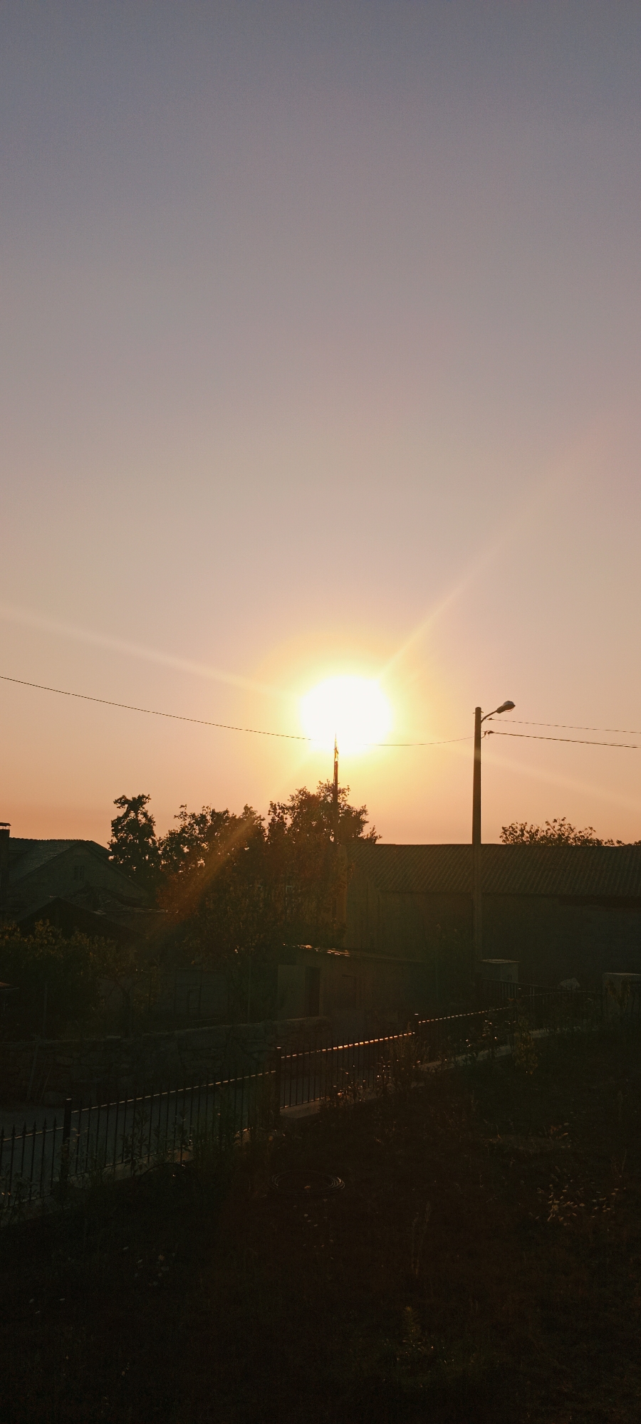 The image depicts a scene at sunset in Adai, Spain. The sky is a gradient of pale purple and soft orange, with the sun positioned prominently in the upper-middle portion of the frame, casting a bright glow and visible rays. A thin power line stretches horizontally across the sky, intersecting the sun's rays. To the right of the sun, a tall utility pole with a light fixture is visible. In the middle ground, a line of trees and various rooftops of buildings are silhouetted against the sky. A fence with vertical bars runs across the lower portion of the image, separating the foreground from the middle ground. The foreground is dark and appears to be overgrown with grass and some plants. The overall atmosphere is that of a calm, late afternoon or early evening in a residential area.