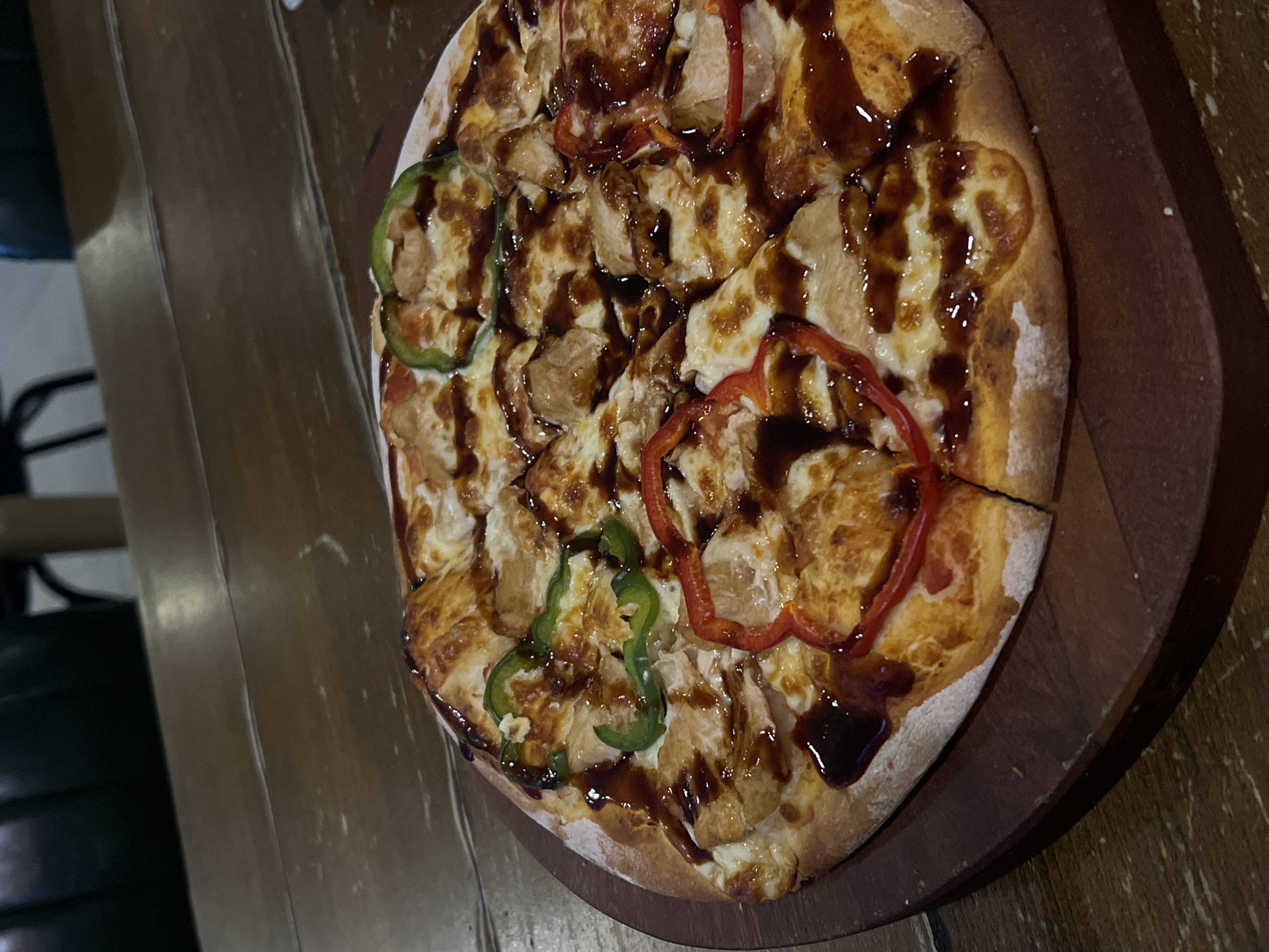 The image shows a pizza on a dark wood-grain surface, likely a table. The pizza features a round crust with a light-colored edge, topped with mozzarella cheese, pieces of what appears to be chicken or another light-colored protein, red and green bell pepper rings, and a dark sauce drizzled over the toppings. The pizza sits on a dark brown wooden plate or tray. The background is blurred, but it includes a glimpse of a black chair frame, and the overall light suggests an indoor restaurant setting. While it is difficult to definitively conclude that the picture was taken in Nha Trang, Vietnam, without more information, there is no conclusive indication that the image could be linked to a specific location.